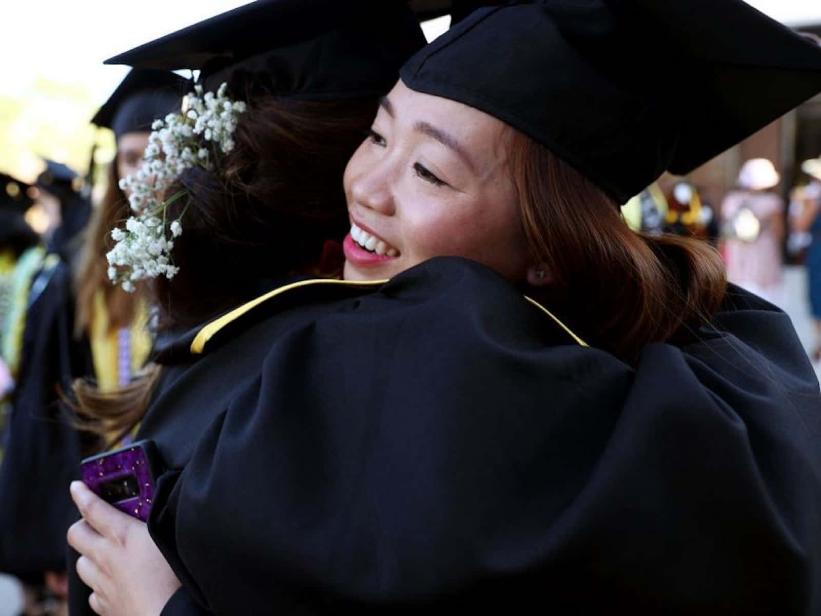 Graduation Ceremony 桌布 壁紙 — 兩位快樂的畢業生身穿黑色學士帽和長袍，頭髮上插滿滿天星，在戶外熱情擁抱。
