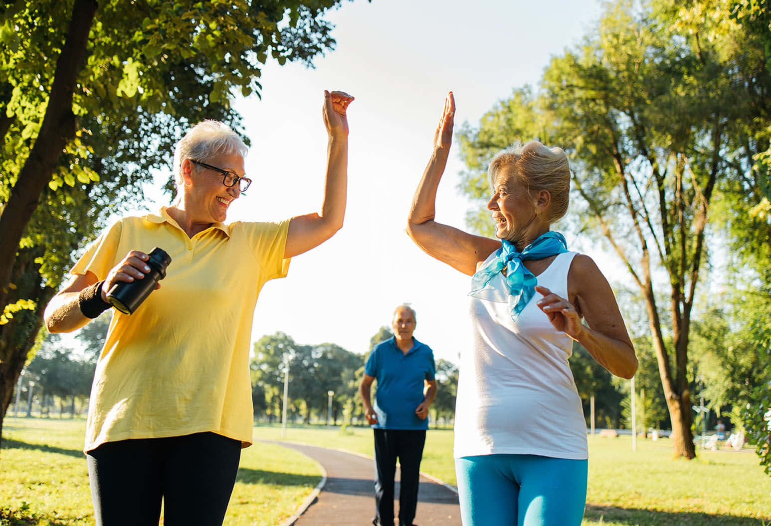Exercise 桌布 壁紙 — 兩位快樂的老年女性在陽光明媚的公園小徑上擊掌，周圍是茂密的綠樹。
