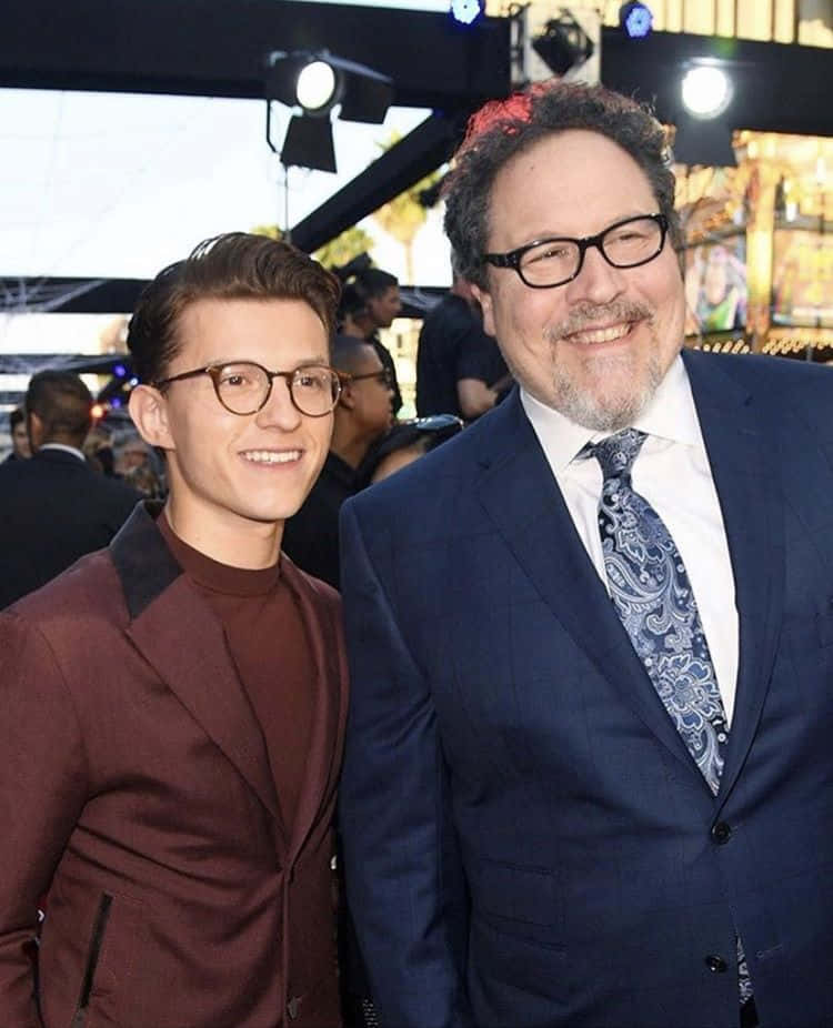 Jon Favreau 桌布 壁紙 — 兩位戴眼鏡的微笑男士在舞台燈光下於明亮活動中一起擺姿勢，背後有模糊的人群。