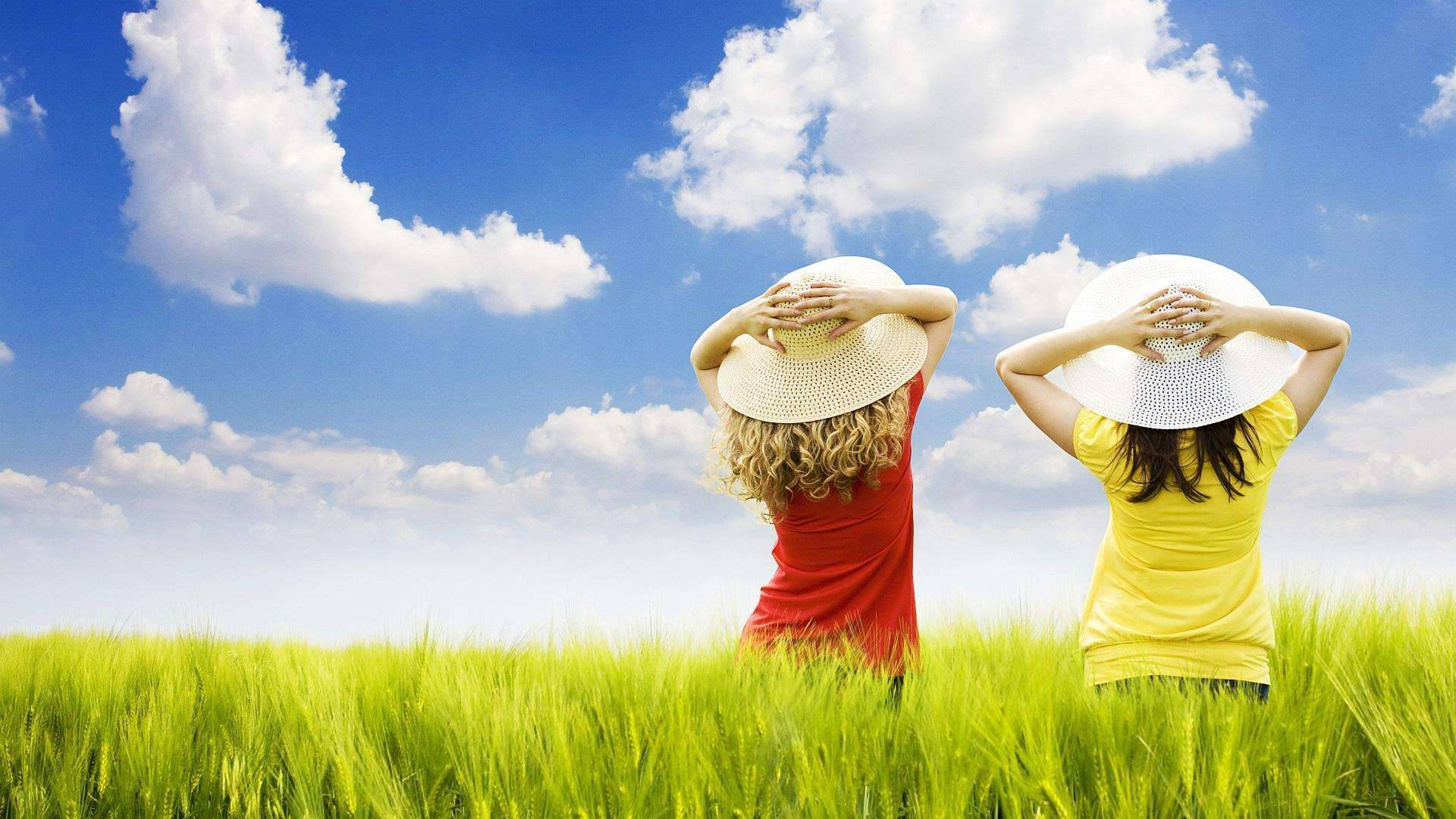 筆電 桌布 壁紙 — 兩位戴草帽的女性在一片充滿活力的綠色田野中背對背站立，頭頂是明亮的藍天和蓬鬆的白雲。