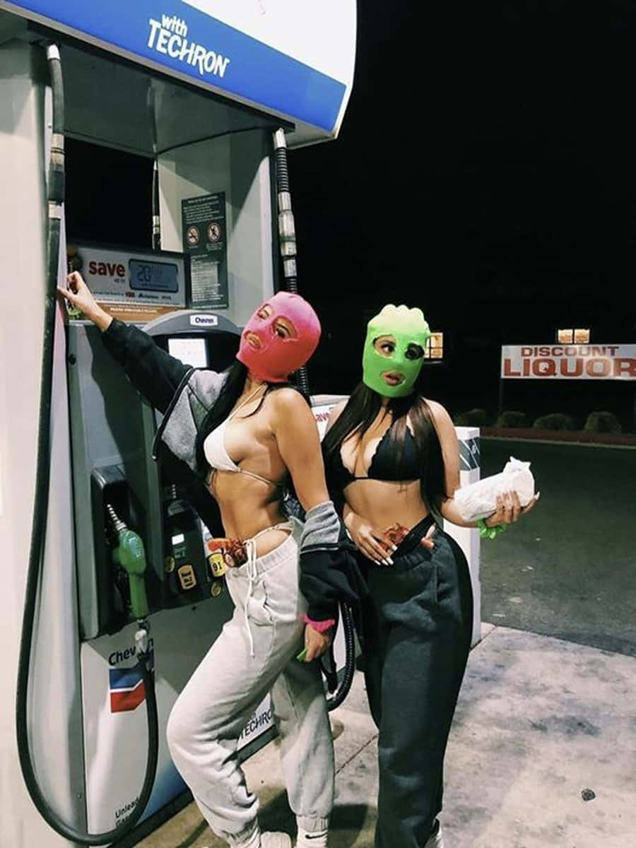 Ski Mask Girl 桌布 壁紙 — 兩位戴著亮粉紅色和綠色口罩的女性在夜燈下的油泵旁擺姿勢，附近有一間折扣酒舖。