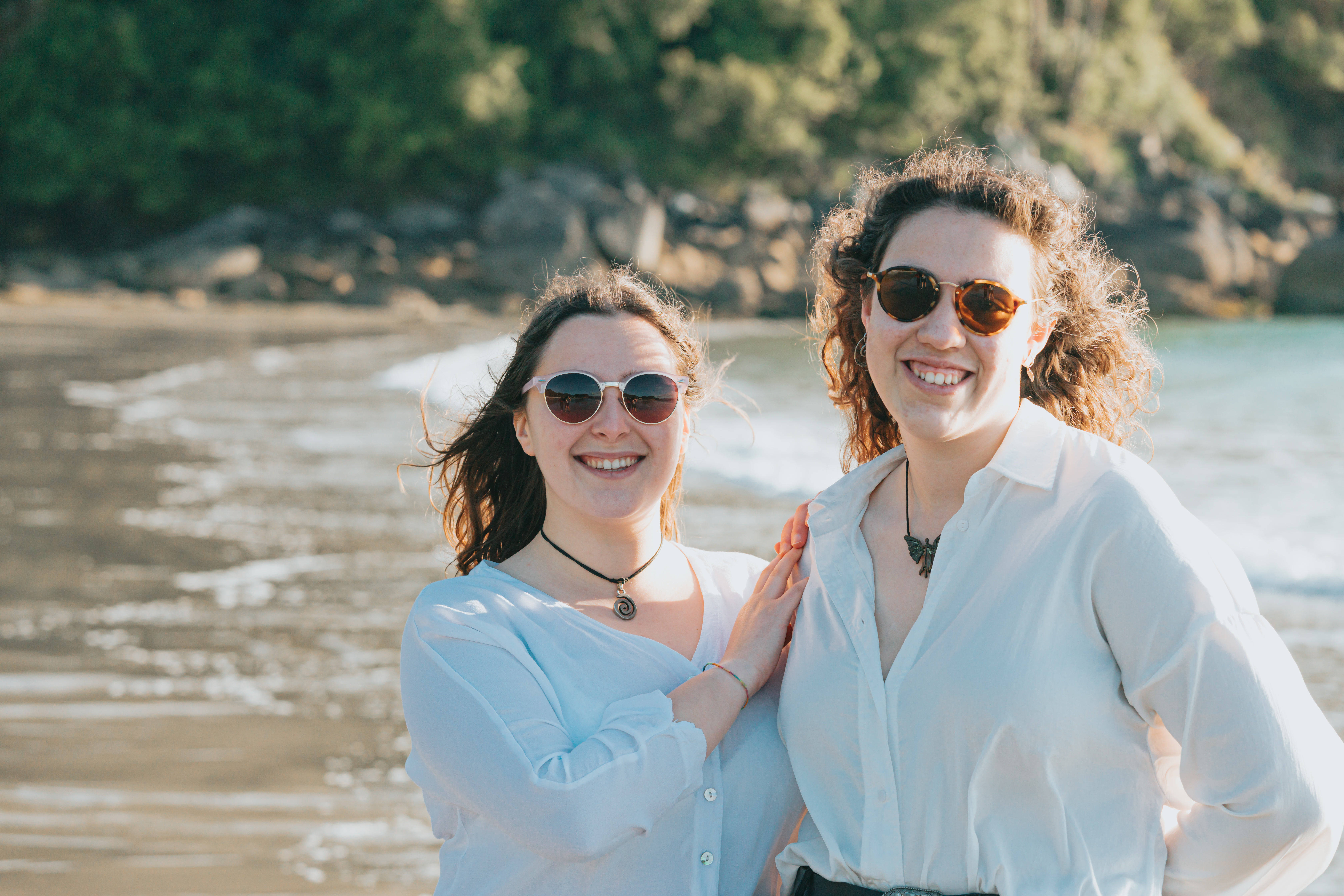 Friendship 桌布 壁紙 — 兩位戴著太陽眼鏡的微笑女士在陽光明媚的海灘上擁抱，身後有輕柔的海浪和綠色懸崖。