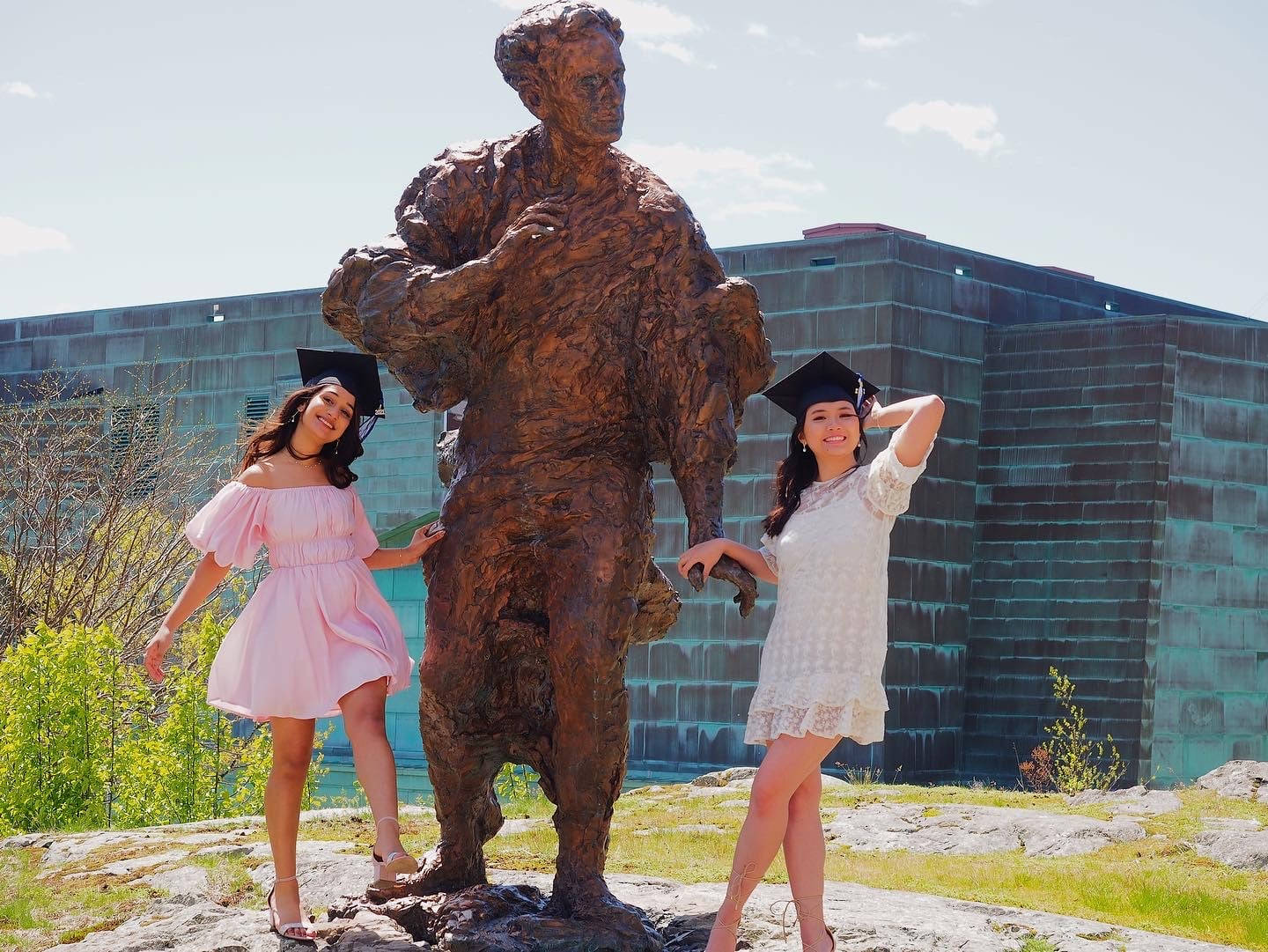 Graduation 桌布 壁紙 — 兩位戴著學士帽穿著畢業袍的畢業生，在陽光明媚的校園日，於一座大型銅像旁俏皮地擺姿勢。