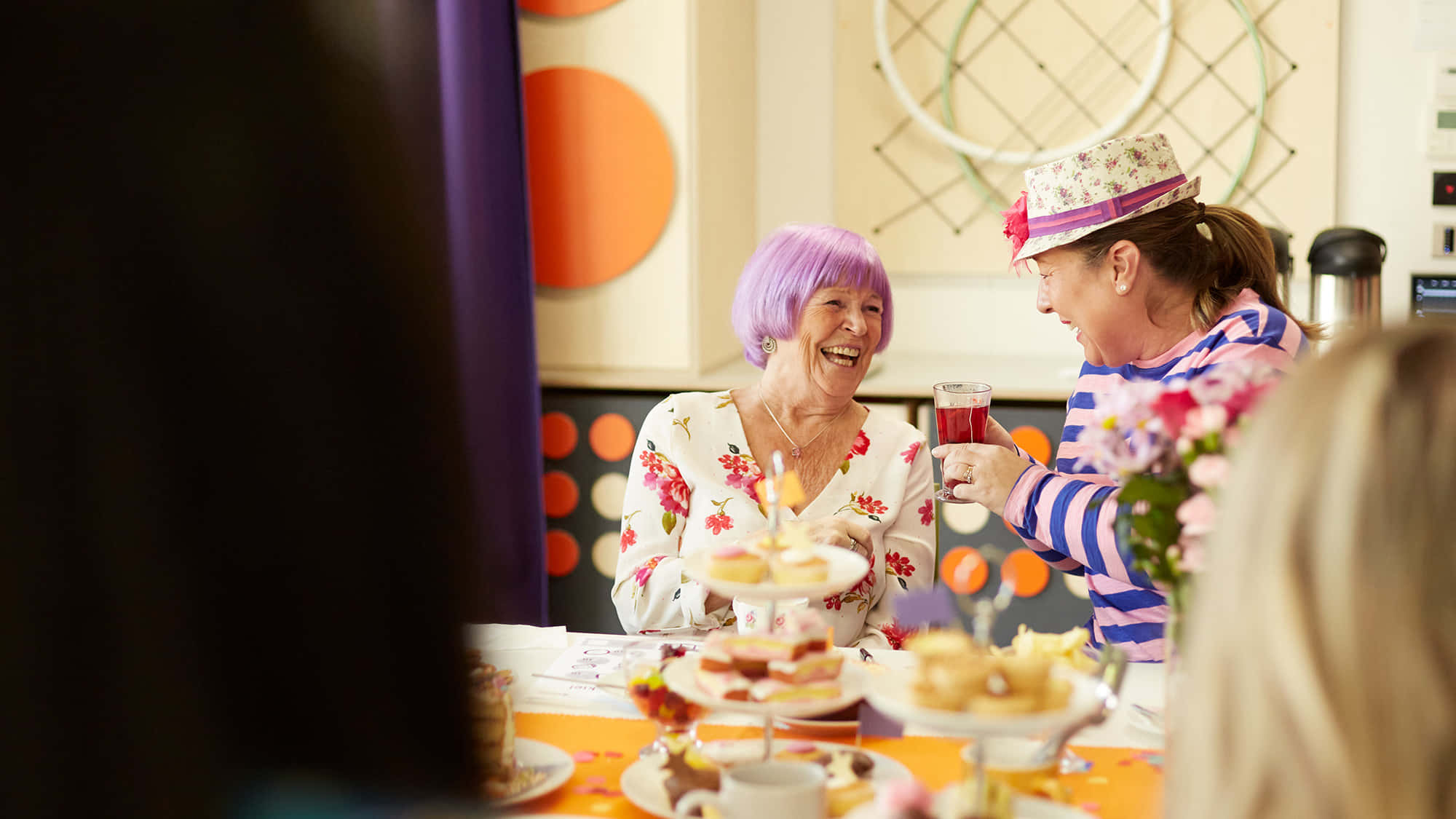 Tea Party 桌布 壁紙 — 兩位戴著派對帽的快樂女性在擺滿蛋糕和美食的彩色桌子上碰杯。
