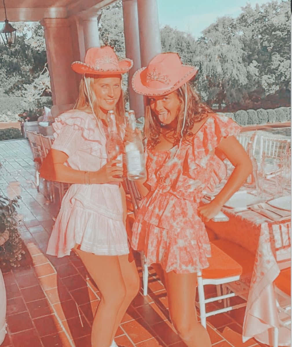 Cowgirl 桌布 壁紙 — 兩位戴著牛仔帽、穿著荷葉邊裙子的女士，在陽光普照、樹木環繞的露臺上，拿著瓶子俏皮地擺姿勢。