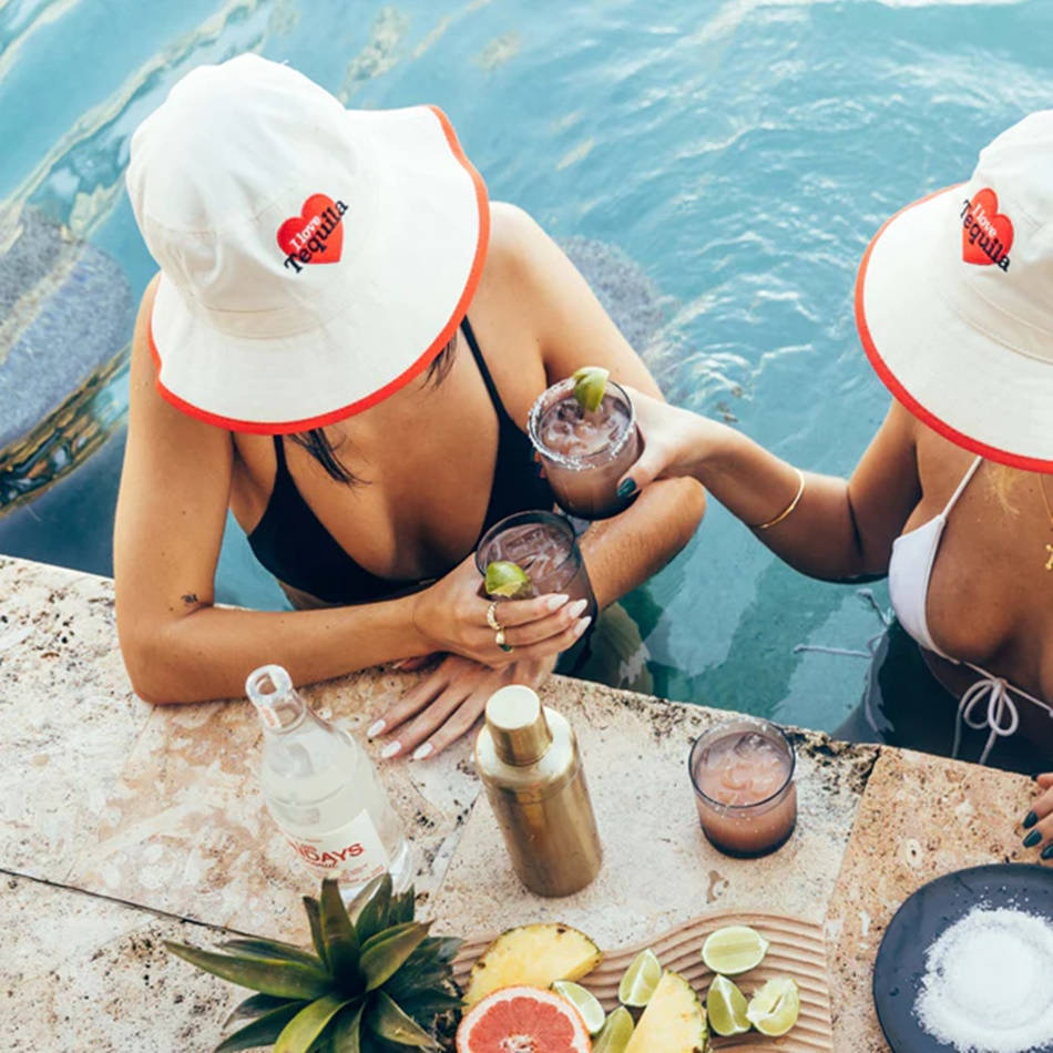 pool 桌布 壁紙 — 兩位戴著白色「I heart tequila」漁夫帽的女性，在泳池邊的酒吧，周圍環繞著熱帶水果和鹽，用瑪格麗特雞尾酒乾杯。