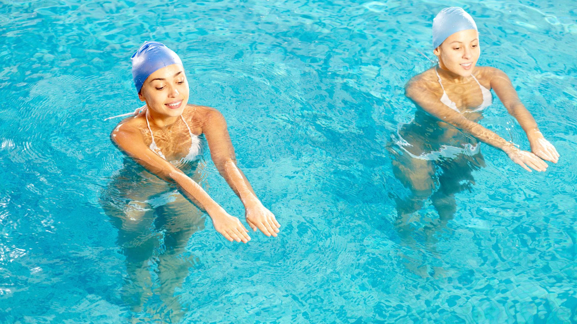 Swimming 桌布 壁紙 — 兩位戴著藍色泳帽的女性，帶著輕鬆的笑容在清澈的碧藍泳池水中滑行。