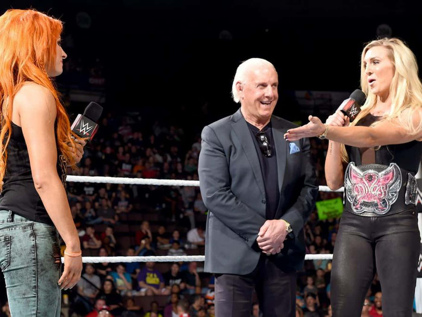 WWE 桌布 壁紙 — 兩位手持麥克風的女WWE摔角手，在歡呼的人群面前，站在摔角擂台內一位西裝笑臉老男人的兩側。