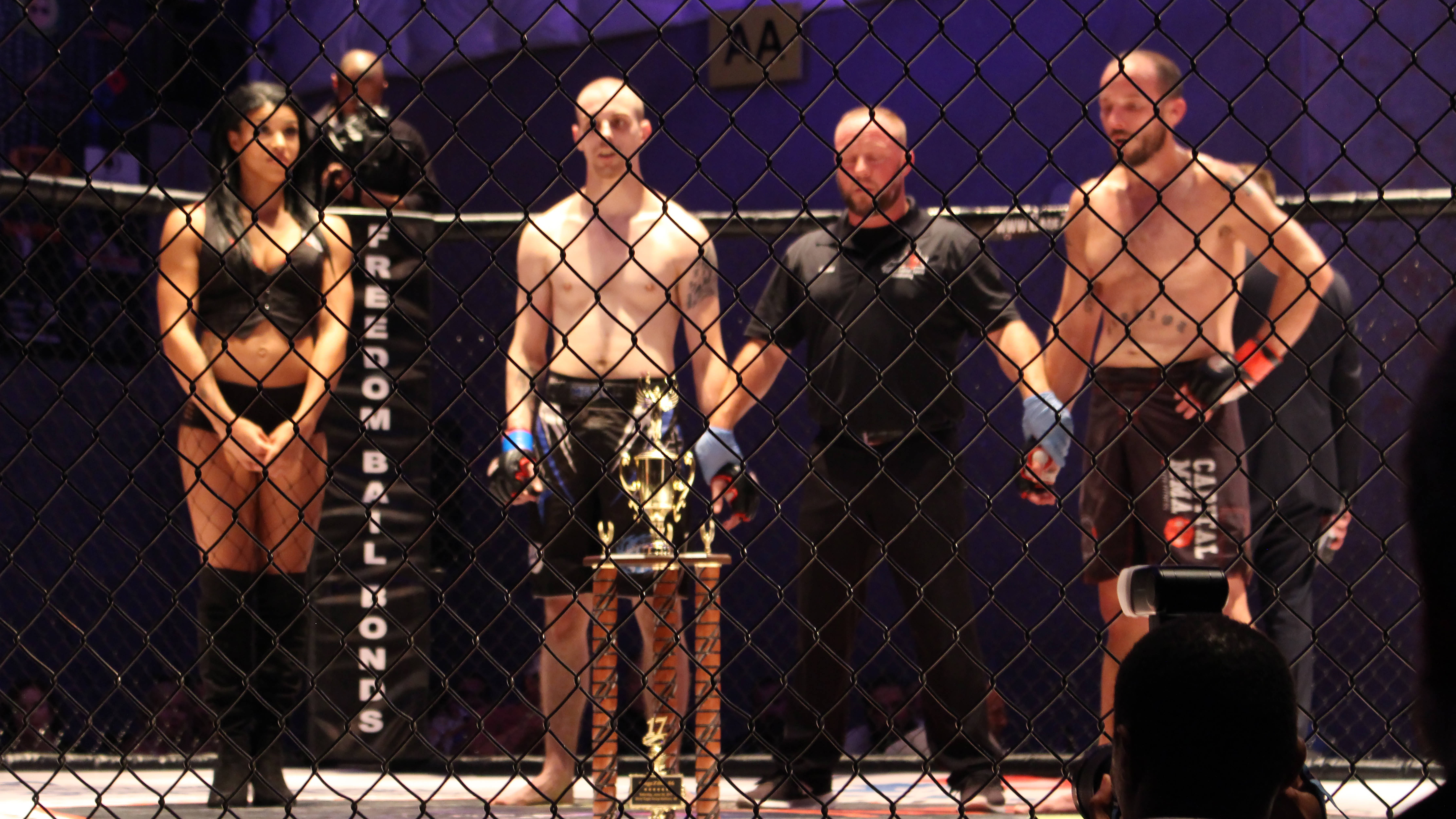 MMA 桌布 壁紙 — 兩位拳手和一位裁判站在一個有獎盃的籠子裡，而一位擂台女郎在綜合格鬥賽事中觀看。
