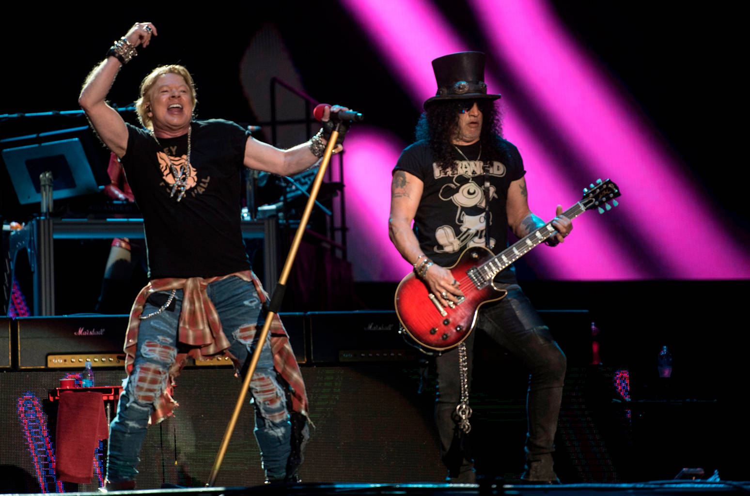 guns n roses 桌布 壁紙 — 兩位搖滾音樂家在充滿活力的粉紅燈光下於舞台上充滿活力地表演。一位高舉雙臂唱歌，另一位戴著高頂帽彈奏紅色電吉他。