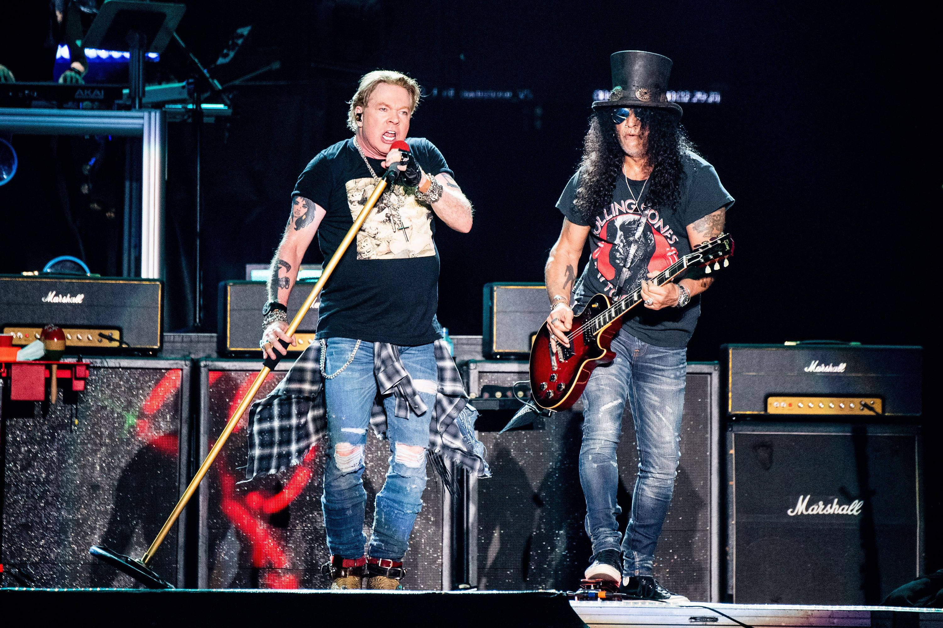 guns n roses 桌布 壁紙 — 兩位搖滾音樂家在明亮的舞台燈光下，手持吉他和麥克風，在舞台上充滿活力地表演。