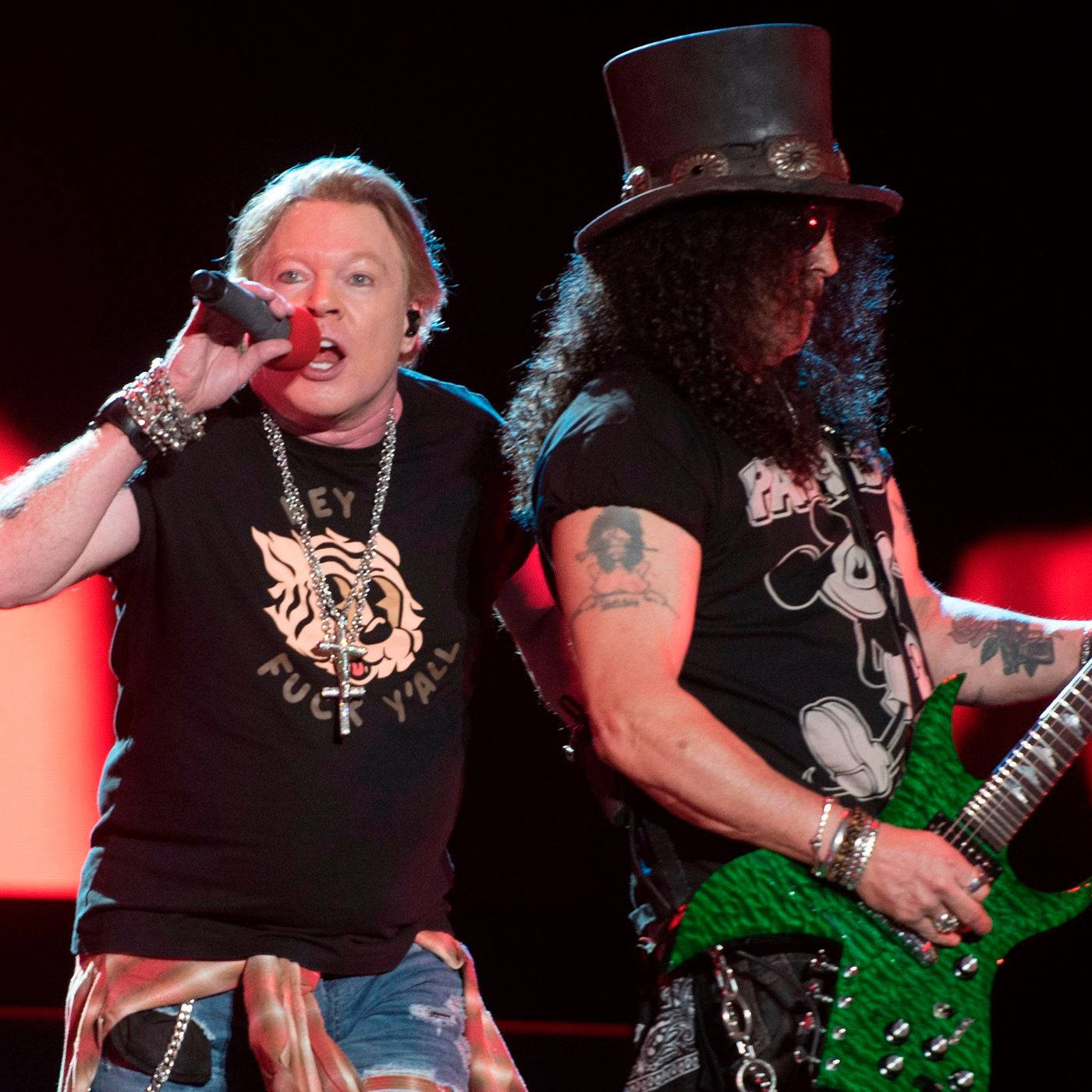 guns n roses 桌布 壁紙 — 兩位搖滾音樂家在舞台上表演，一位對著麥克風唱歌，另一位在紅色舞台燈光下彈奏綠色電吉他。