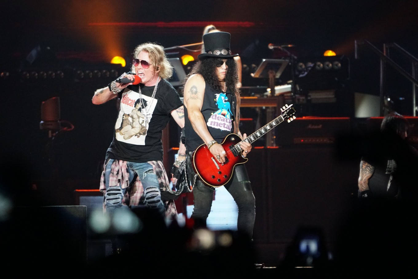 rock and roll 桌布 壁紙 — 兩位搖滾音樂家在舞台上表演，一位對著麥克風唱歌，另一位在明亮的舞台燈光下彈奏紅色電吉他。