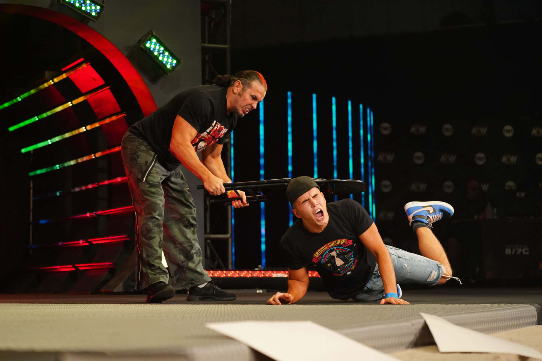 WWE 桌布 壁紙 — 兩位摔角手在戲劇性的比賽中，一人手持鋼椅舉過另一人頭頂，後者跪在舞台上。