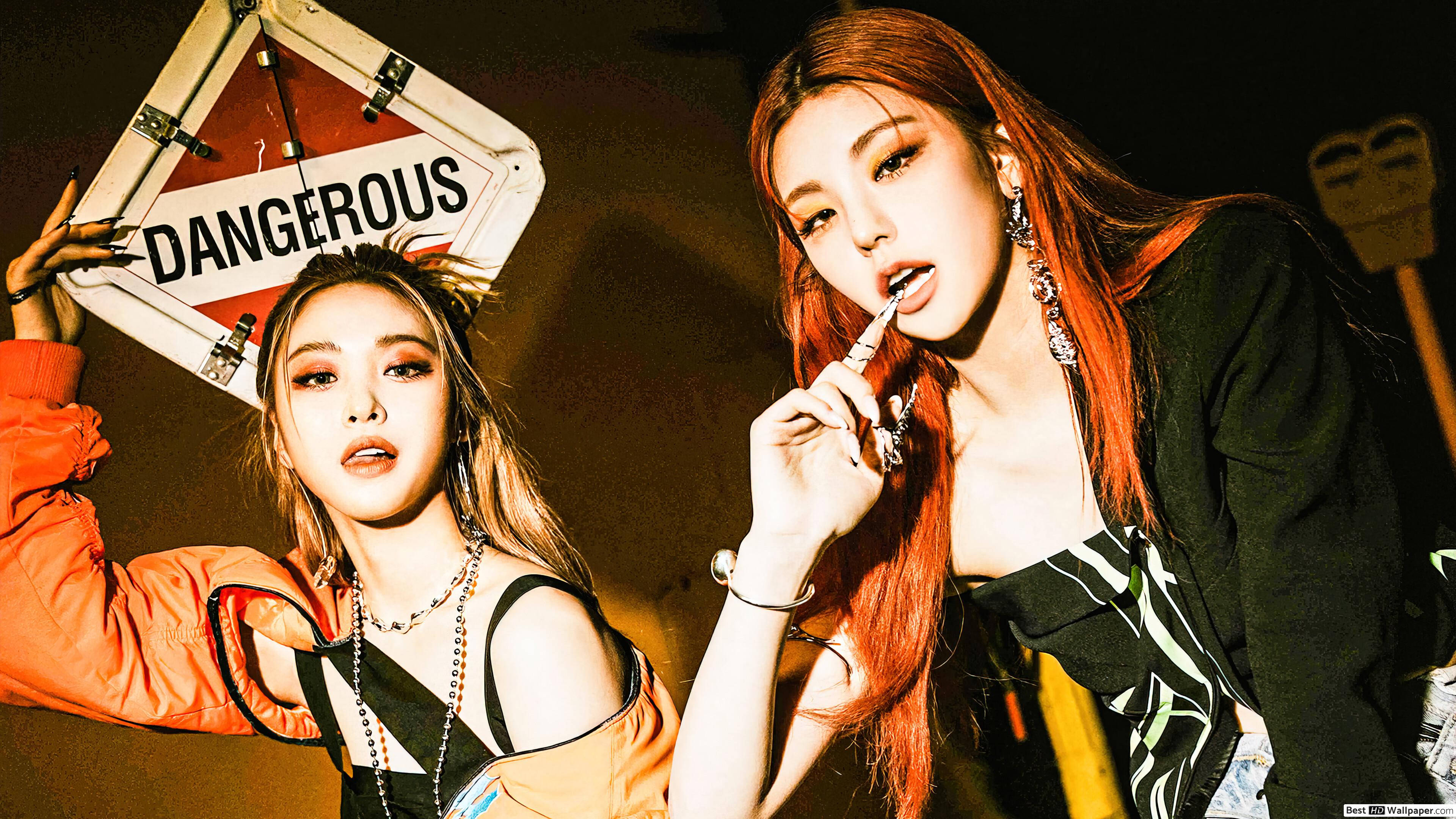 ITZY 桌布 壁紙 — 兩位時尚女性在夜晚戲劇性地擺姿勢，一人手持「DANGEROUS」標誌，另一人在溫暖燈光下舔唇膏管。