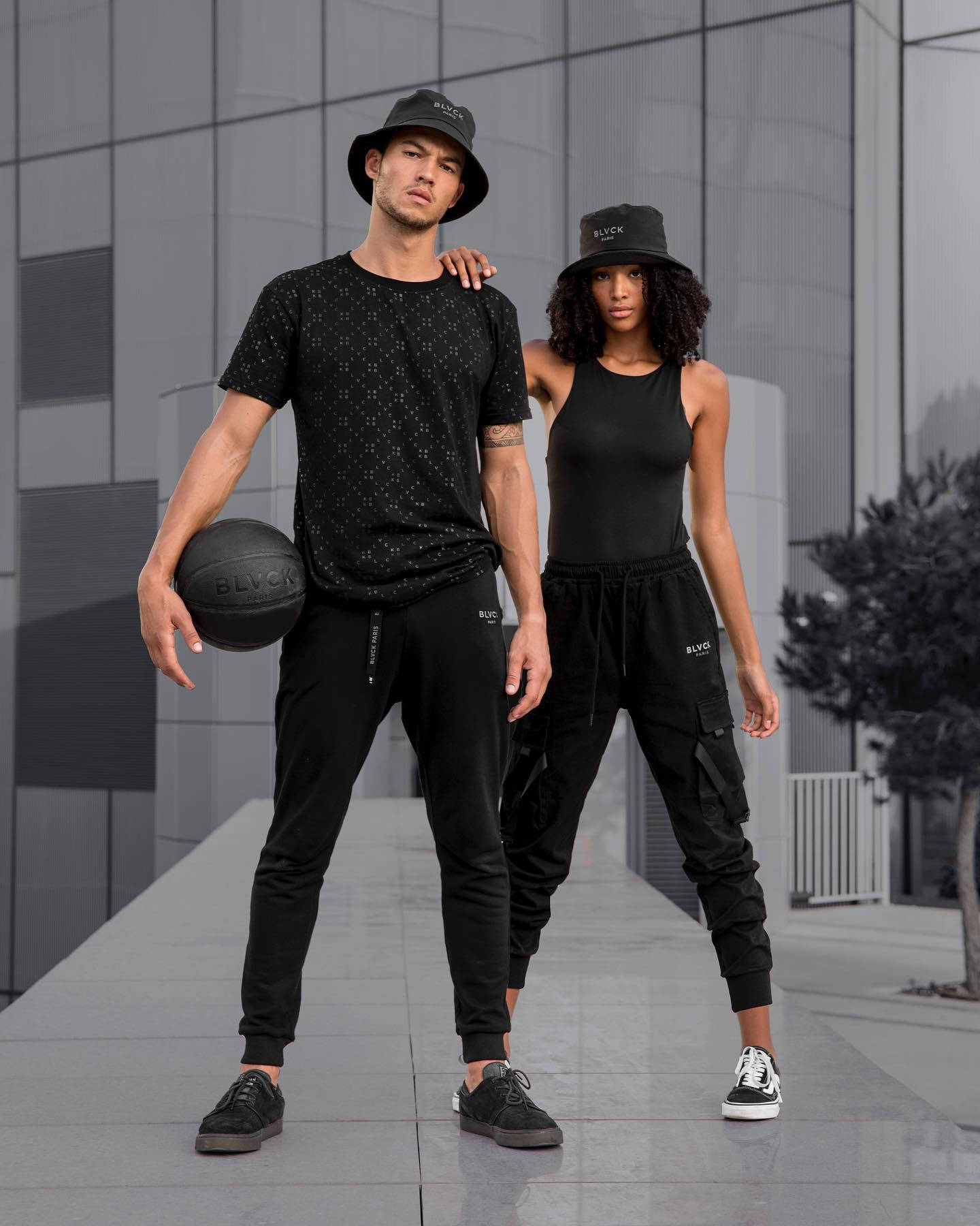 clothing 桌布 壁紙 — 兩位時尚模特兒身穿全黑運動服，自信地拿著籃球，以現代玻璃建築外牆為背景擺姿勢。