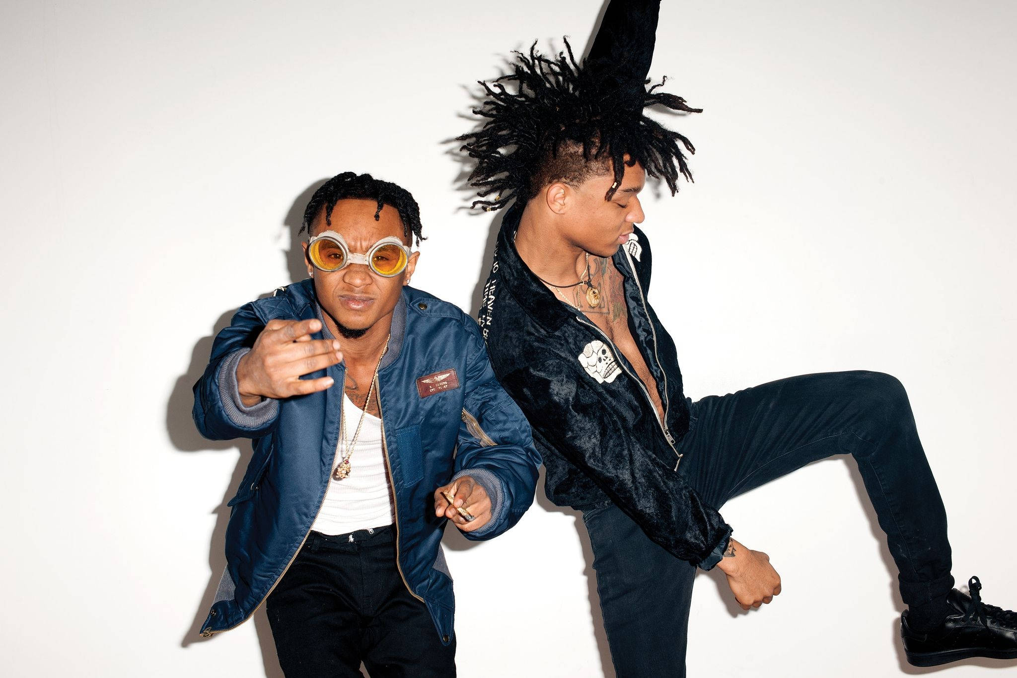 Swae Lee 桌布 壁紙 — 兩位時尚男士在純白背景下動感擺姿勢，一位身穿藍色外套戴圓形太陽眼鏡，另一位身穿黑色天鵝絨外套，頭髮狂野。