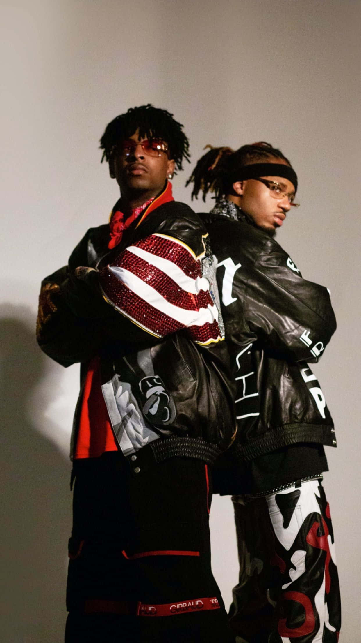 21 Savage 桌布 壁紙 — 兩位時尚男士穿著大膽的黑色皮夾克，搭配紅、白、黑的圖形設計，在簡約的灰色工作室牆前自信地擺姿勢。