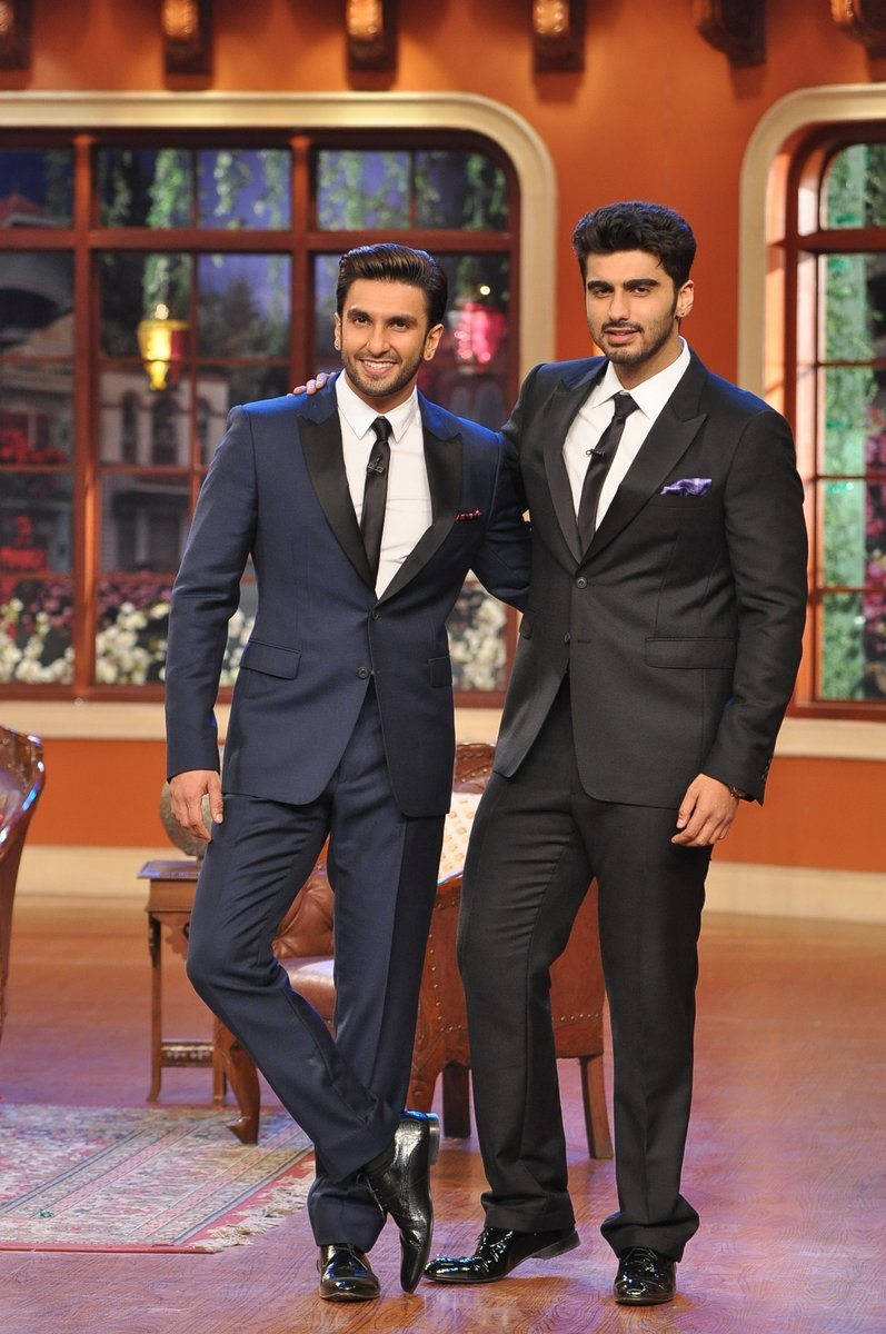 Arjun Kapoor 桌布 壁紙 — 兩位時尚男士身穿筆挺西裝，在燈光溫暖的脫口秀場景中自信擺姿勢，背景有華麗窗戶和復古家具。