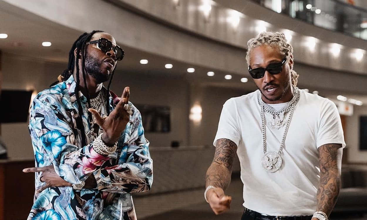 2 Chainz 桌布 壁紙 — 兩位時尚的饒舌歌手戴著太陽眼鏡，在溫暖燈光的現代室內大廳自信地做手勢。