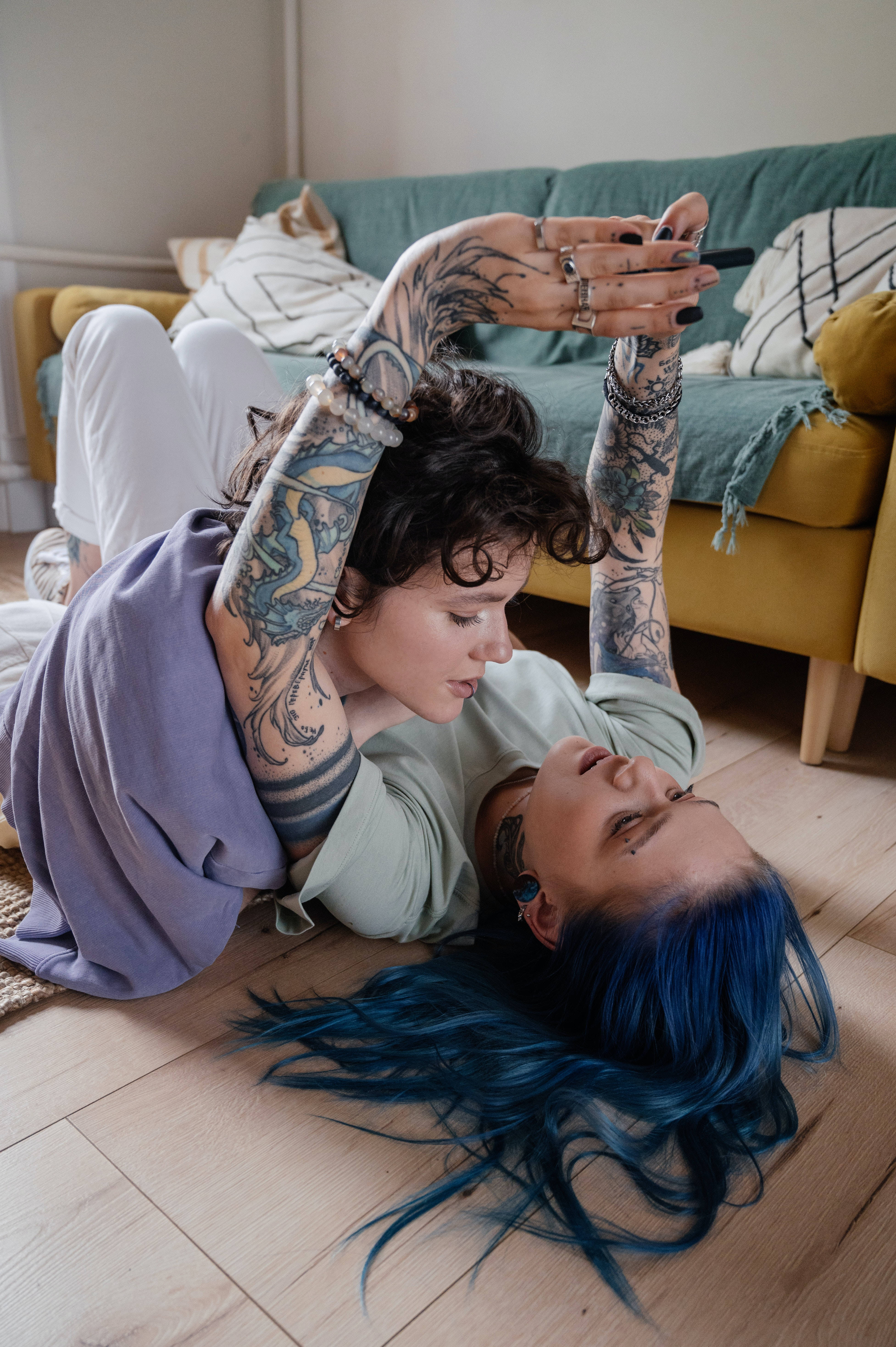 Tattoo 桌布 壁紙 — 兩位有刺青的藍髮女士一起躺在木地板上，在舒適的客廳裡使用手機。