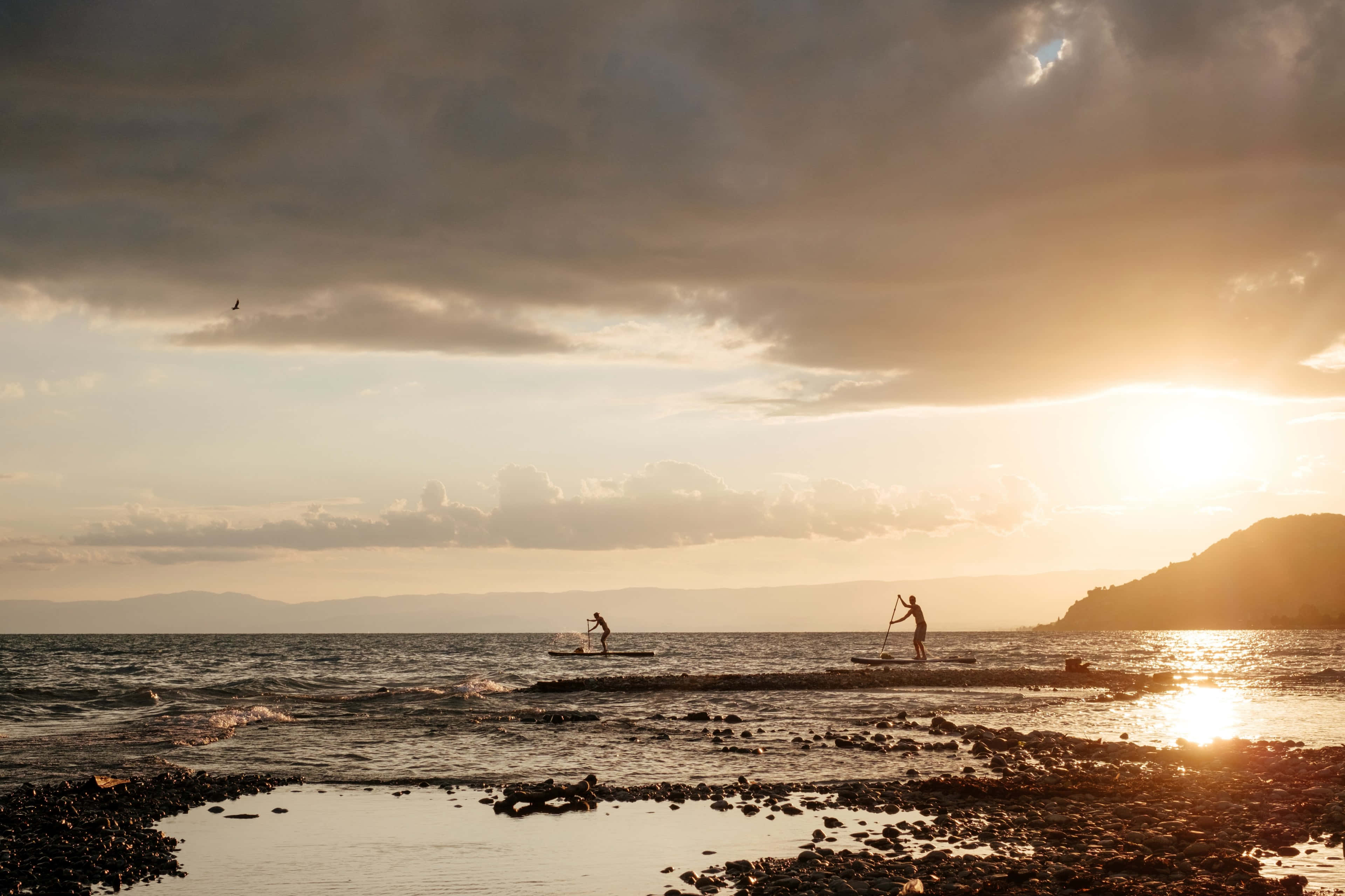 Paddleboarding 桌布 壁紙 — 兩位槳板運動員在日落時分滑過平靜的海面，金色光芒反射在水面上，上方有烏雲。