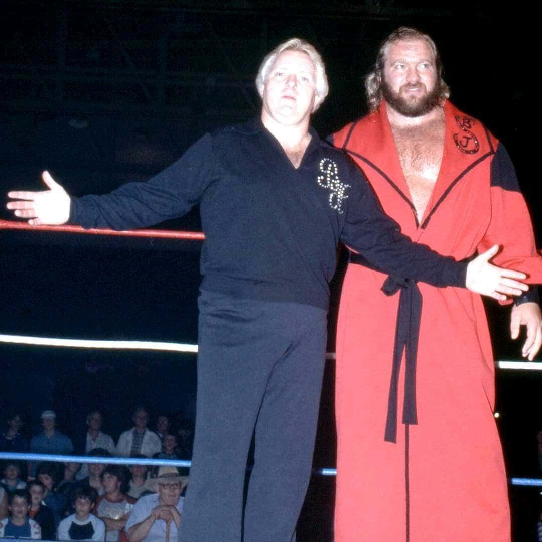 Bobby Heenan 桌布 壁紙 — 兩位標誌性摔角手站在擂台上，一位身穿黑色服裝，另一位身穿紅色長袍，在明亮的場館燈光下向觀眾做手勢。