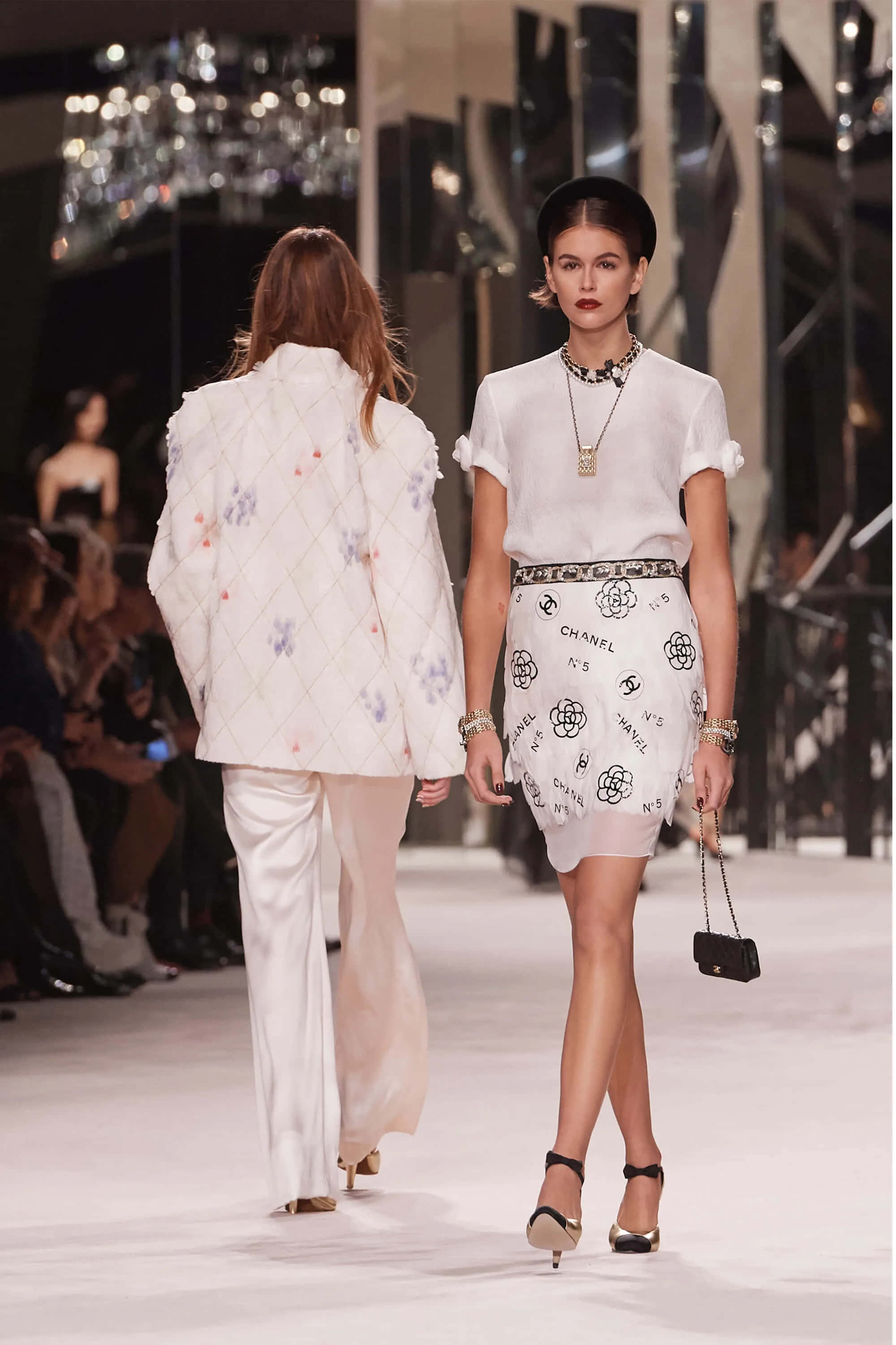 Clothes 桌布 壁紙 — 兩位模特兒在明亮的表演燈光下，穿著優雅的白色 Chanel 服裝，在閃亮的 T 台上大步走著。