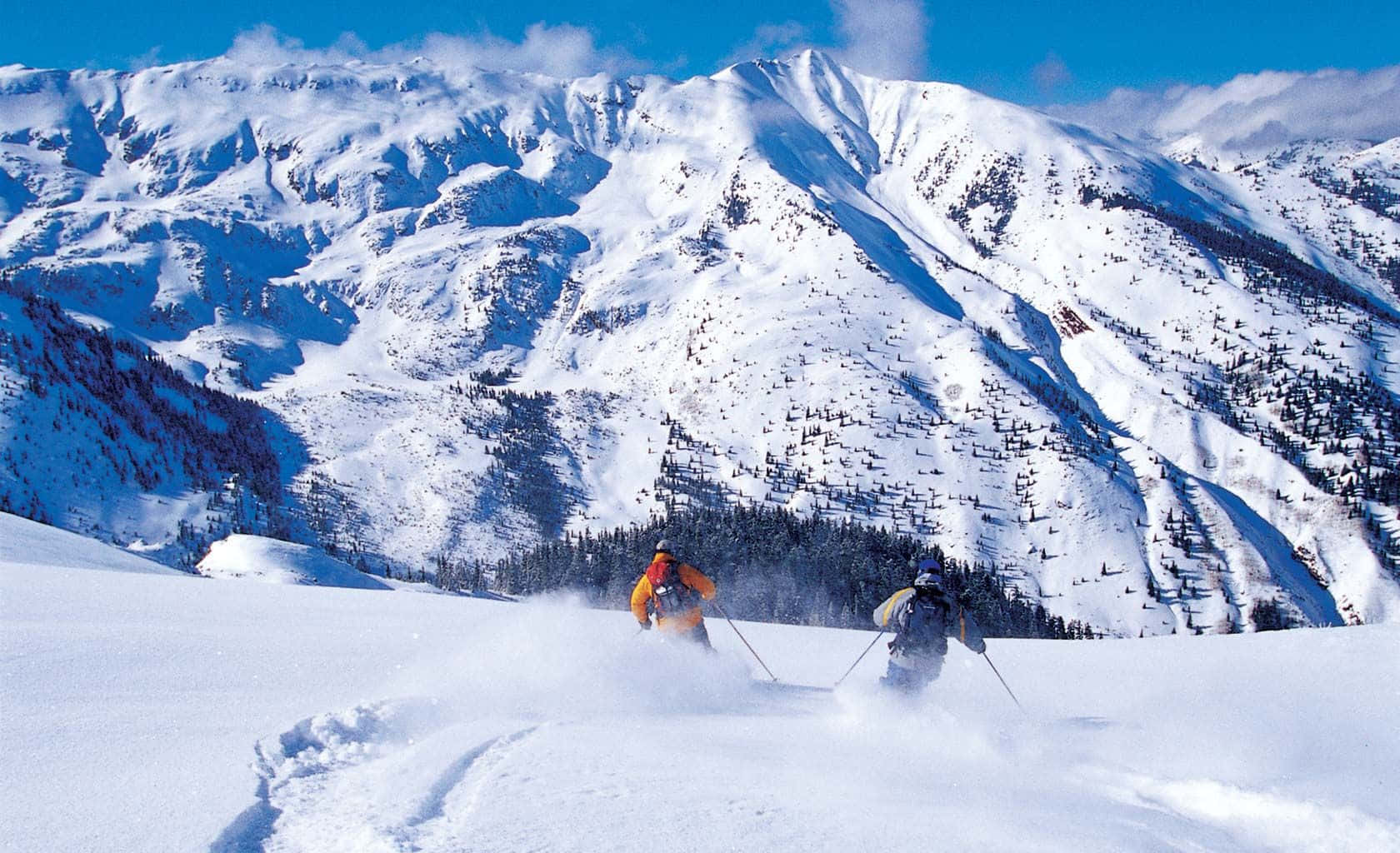Aspen Colorado 桌布 壁紙 — 兩位滑雪者在陽光照耀的雪坡上劃過新鮮的粉雪，背後聳立著崎嶇的山峰。