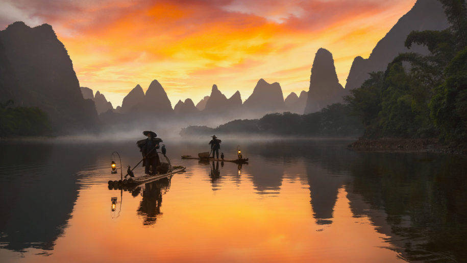 Beijing 桌布 壁紙 — 兩位漁夫在日落時分乘竹筏滑過迷霧水域，喀斯特山脈在火紅橙雲的映襯下形成剪影。
