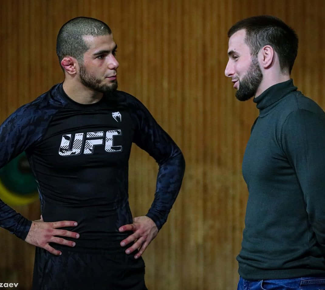 MMA 桌布 壁紙 — 兩位激烈的男性拳手在木牆健身房中對峙，一人身穿UFC襯衫，另一人穿著深色高領衫。