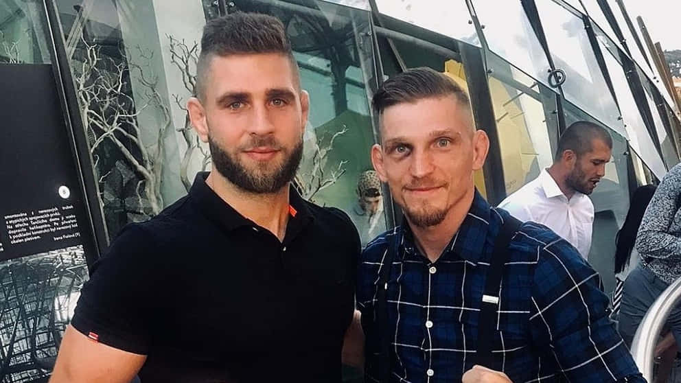 MMA 桌布 壁紙 — 兩位留著時尚髮型和鬍子的男士，在映有樹木倒影的現代玻璃建築前合影。