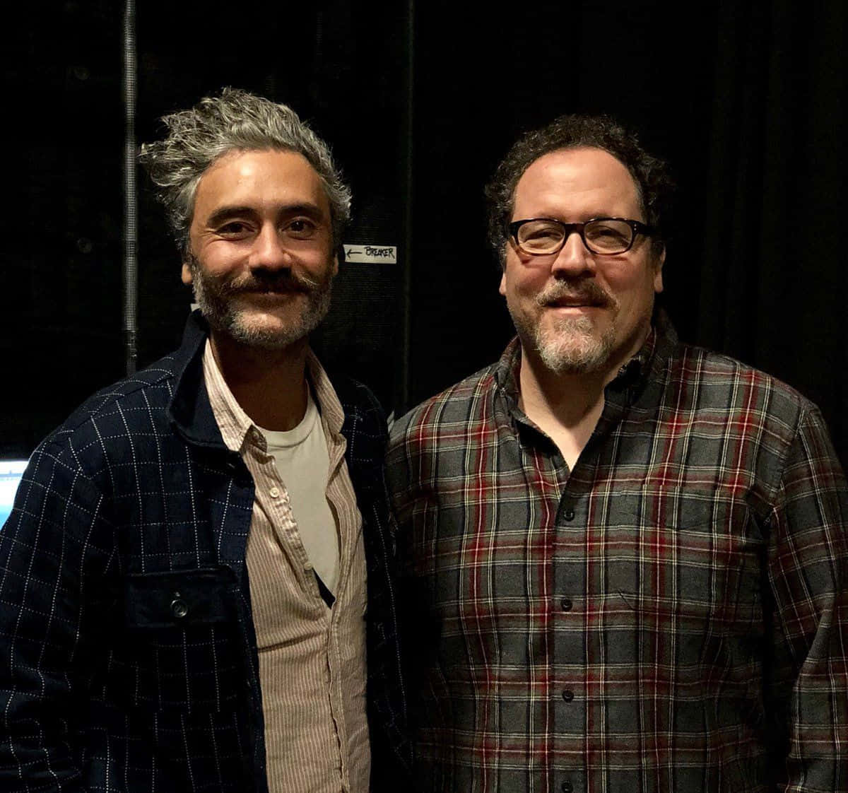 Jon Favreau 桌布 壁紙 — 兩位留鬍鬚、身穿格紋襯衫的男士在黑色布幕後台一起擺姿勢微笑。