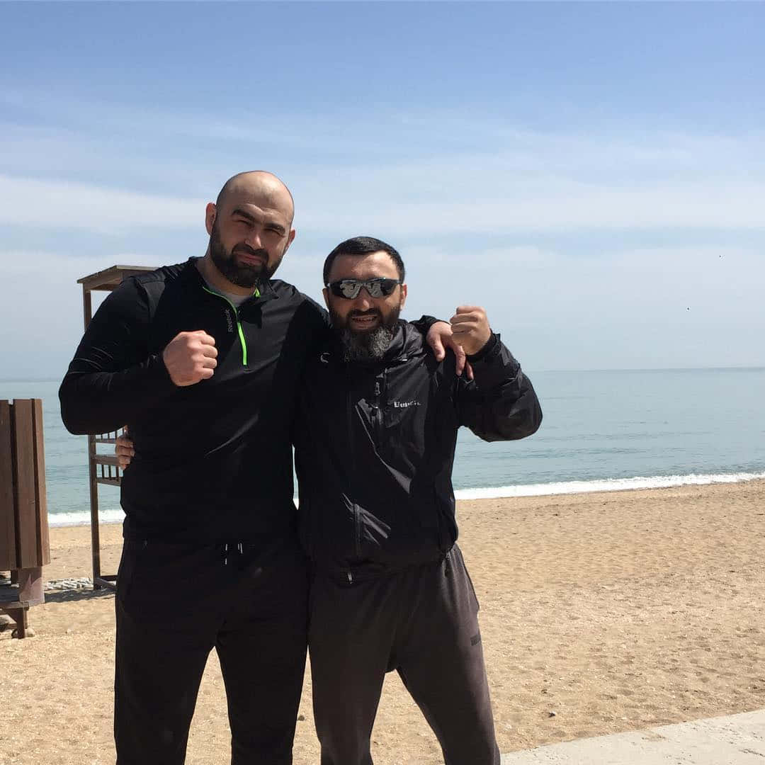 MMA 桌布 壁紙 — 兩位留鬍鬚的男士身穿黑色運動服，在陽光明媚的海灘上自信地擺姿勢，背景是海浪和清澈的藍天。