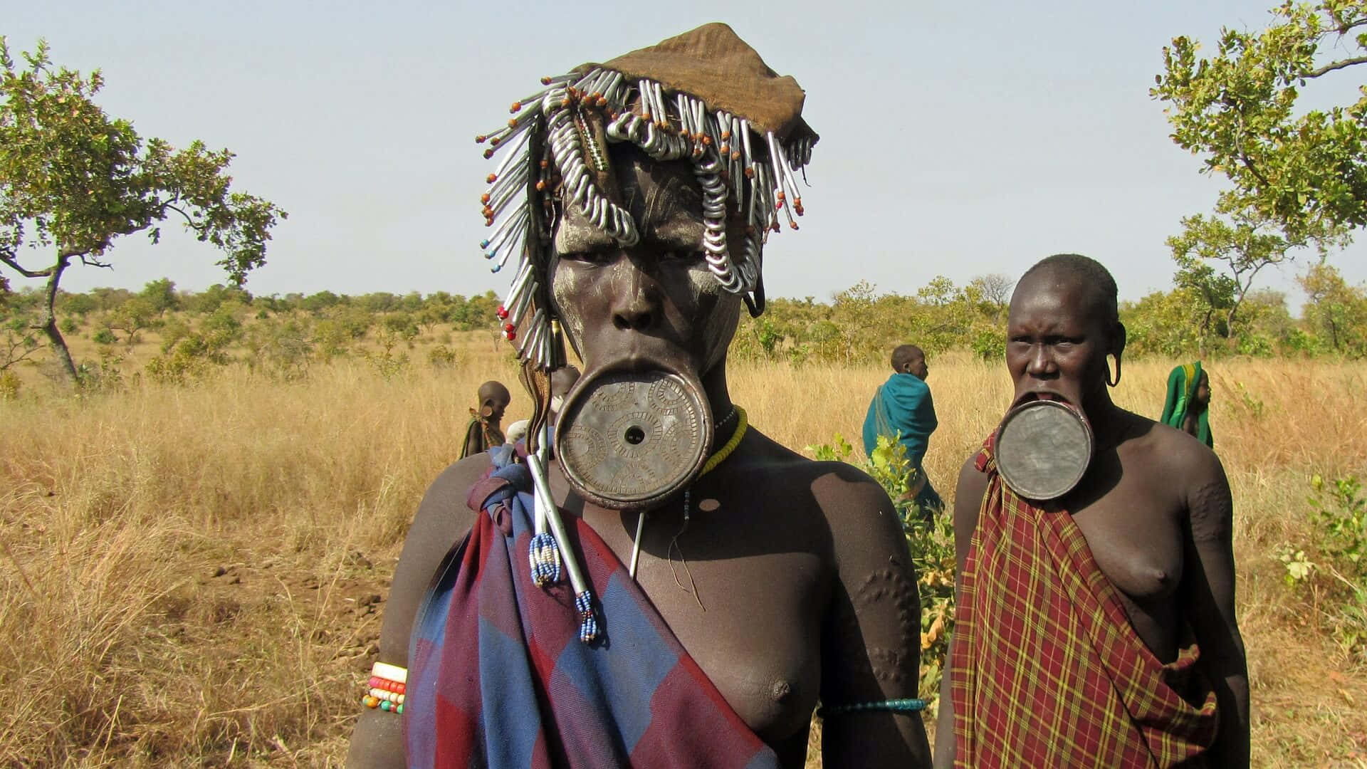 African 桌布 壁紙 — 兩位穆爾西族女性，戴著大型唇盤和珠飾頭飾，站在晴朗天空下的乾燥草原稀樹草原上。