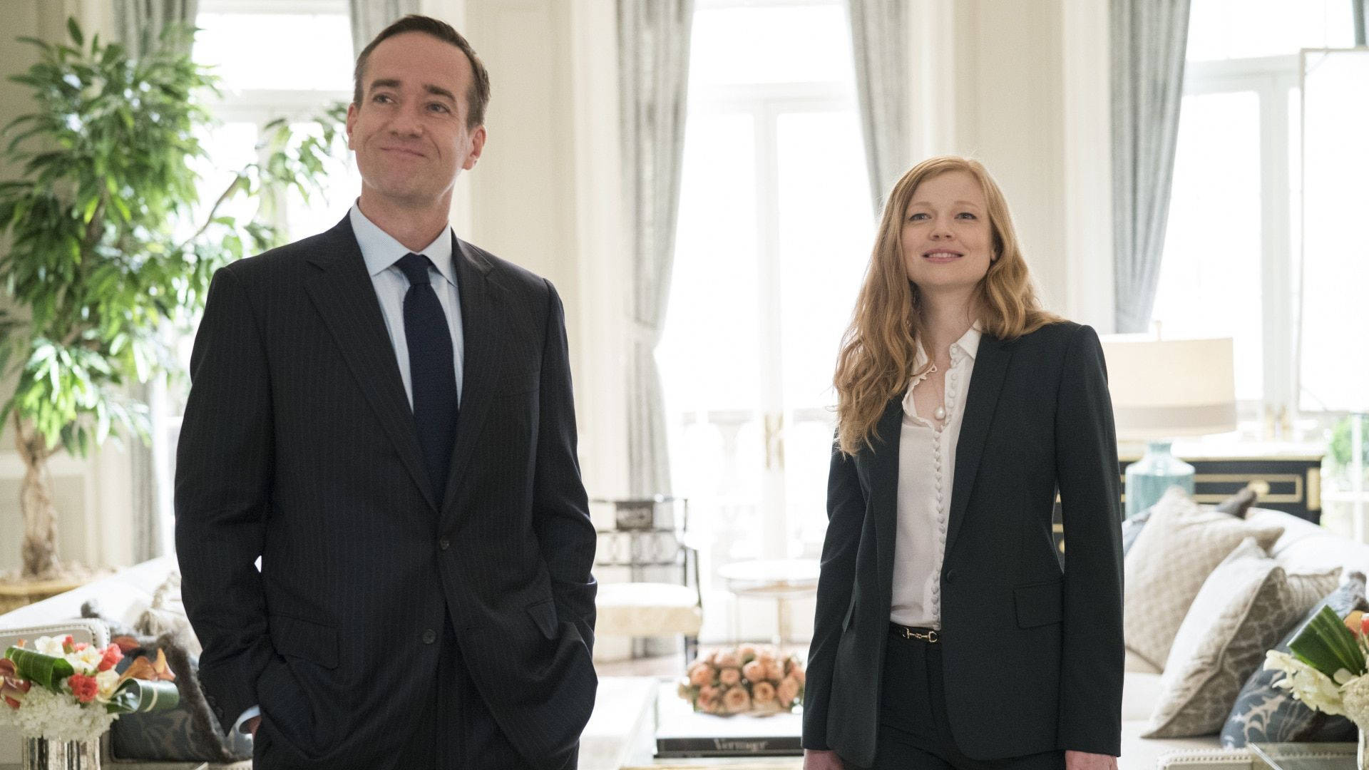 Succession 桌布 壁紙 — 兩位穿著正式服裝的人士站在一個光線充足、裝潢奢華的客廳中，客廳有大窗戶和優雅的裝飾。