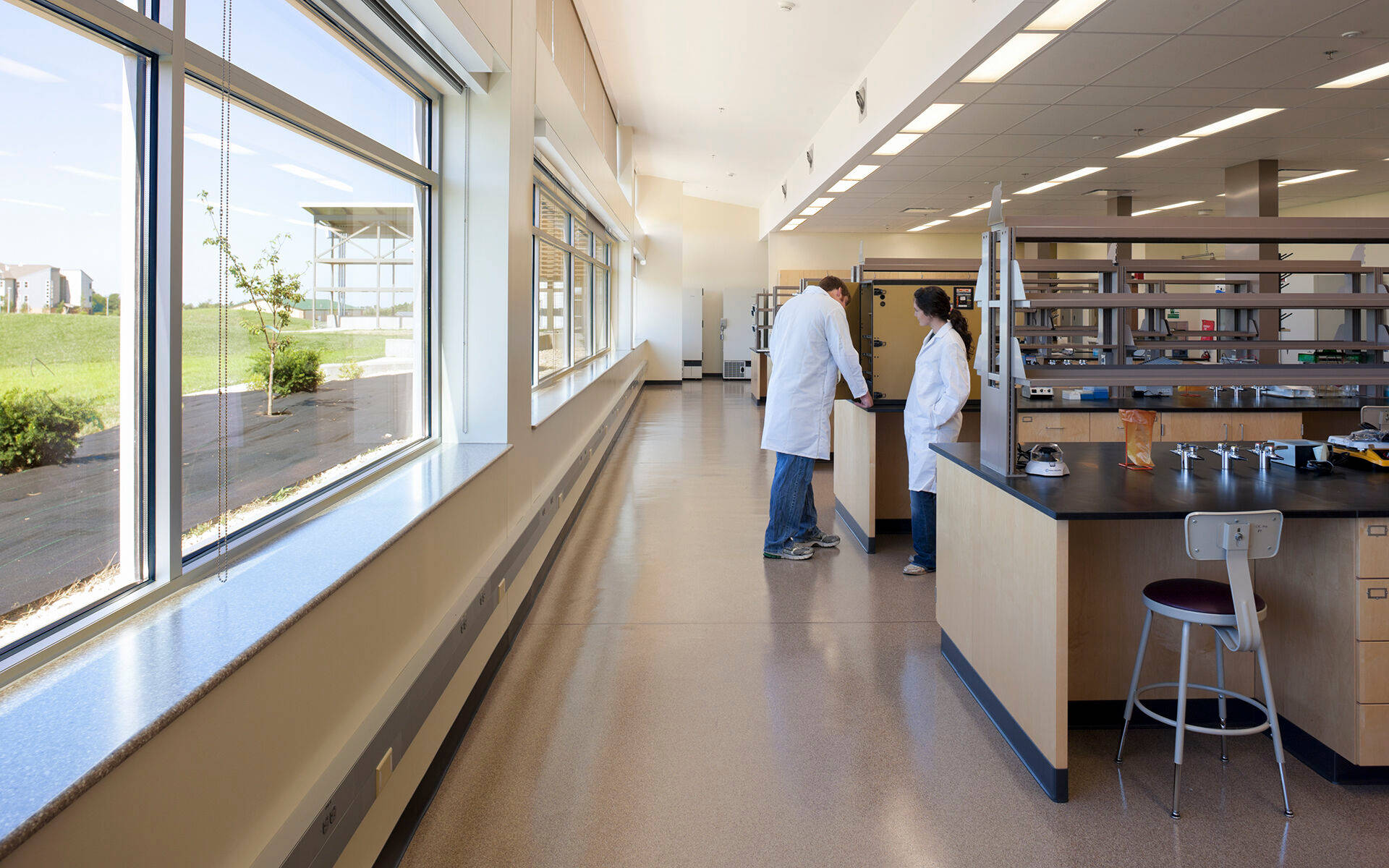 Laboratory 桌布 壁紙 — 兩位穿著白色實驗袍的科學家在明亮的燈光下於實驗桌工作，大窗戶顯示外面的綠色草地。