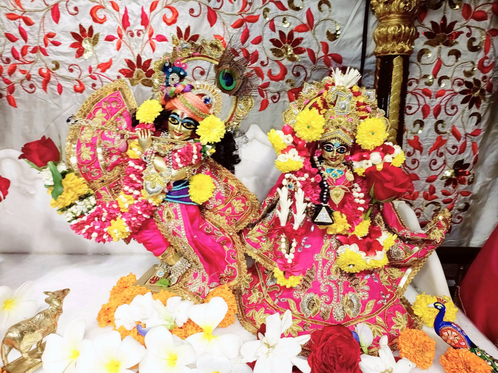 radha krishna HD 桌布 壁紙 — 兩位穿著華麗的印度教神祇，飾以粉紅、金色和黃色花朵，坐在紅色花卉刺繡背景前。