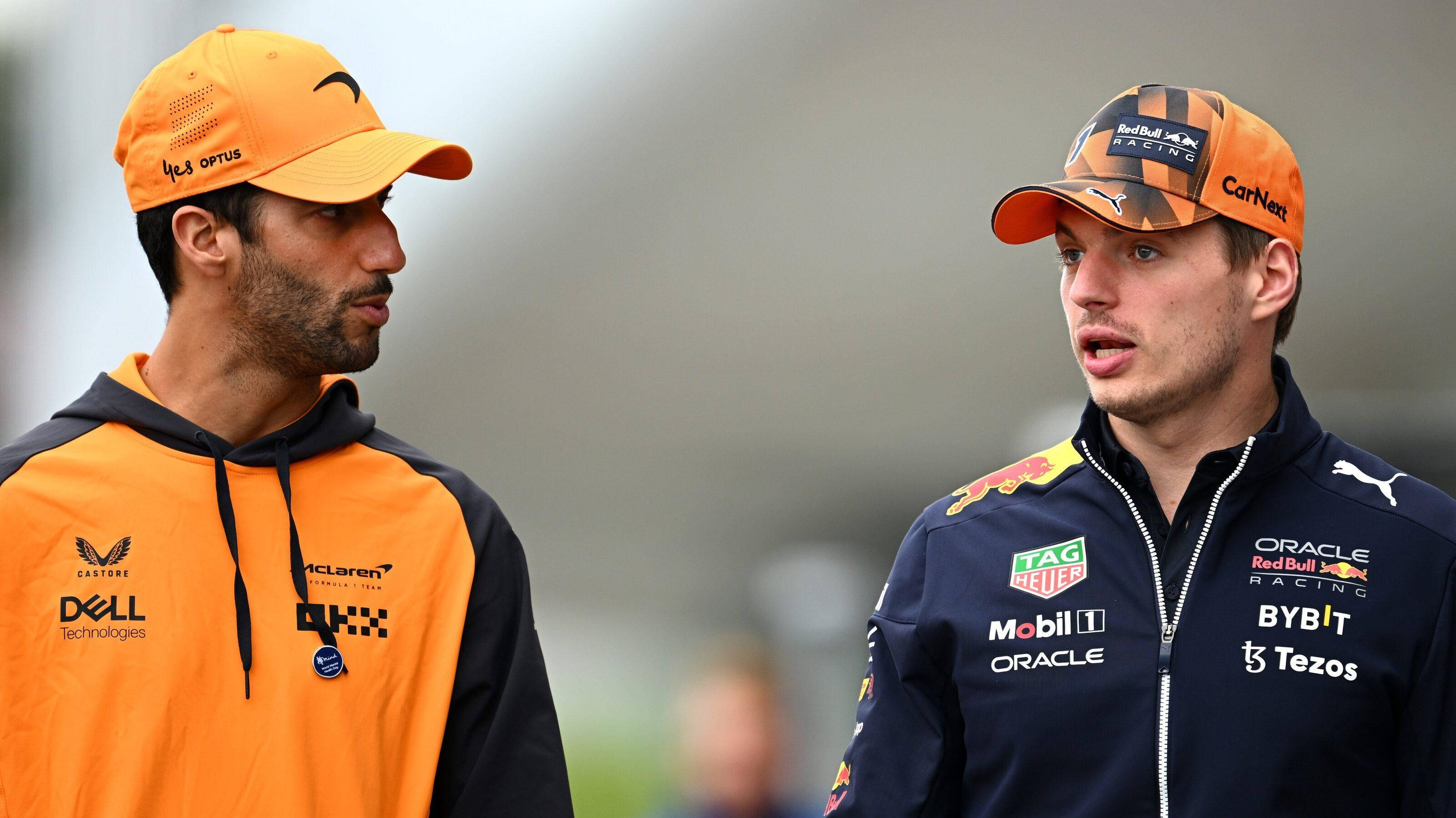 Max Verstappen 桌布 壁紙 — 兩位穿著車隊裝備的F1車手在模糊背景的比賽週末中專注交談。