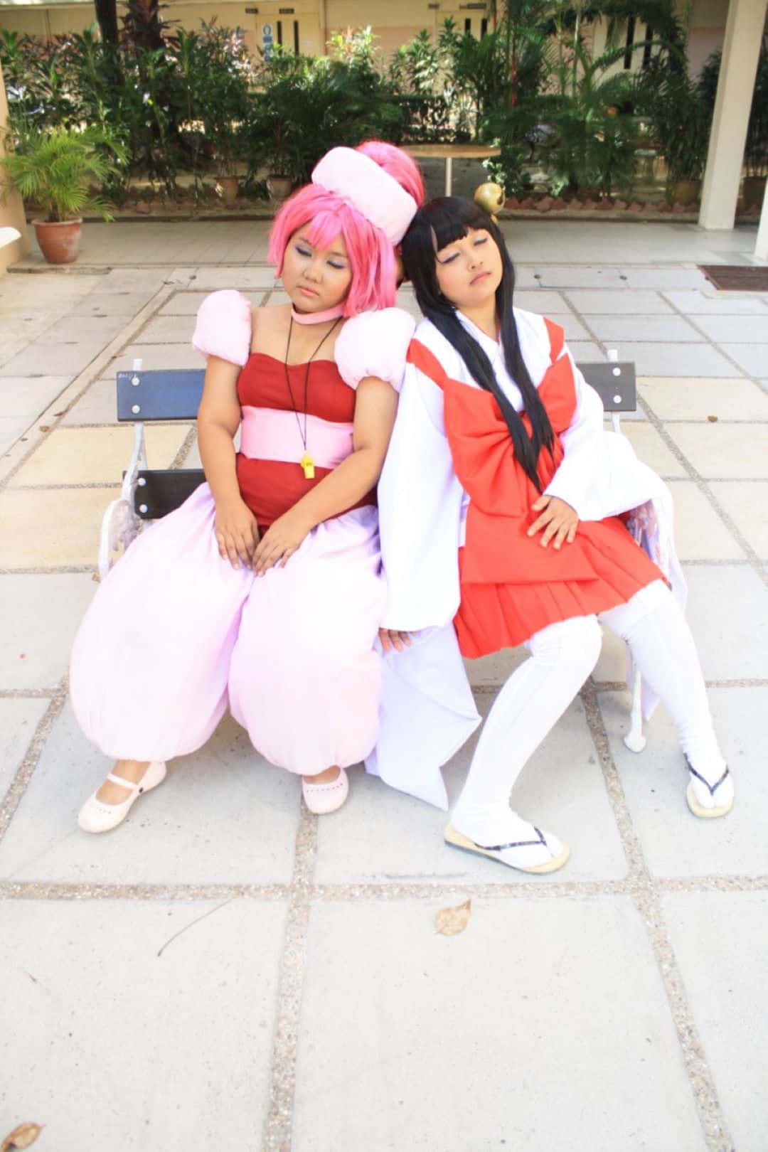  壁紙 — 兩位穿著鮮豔動漫服裝的cosplayer並排坐在陽光普照、綠意盎然的庭院長椅上。