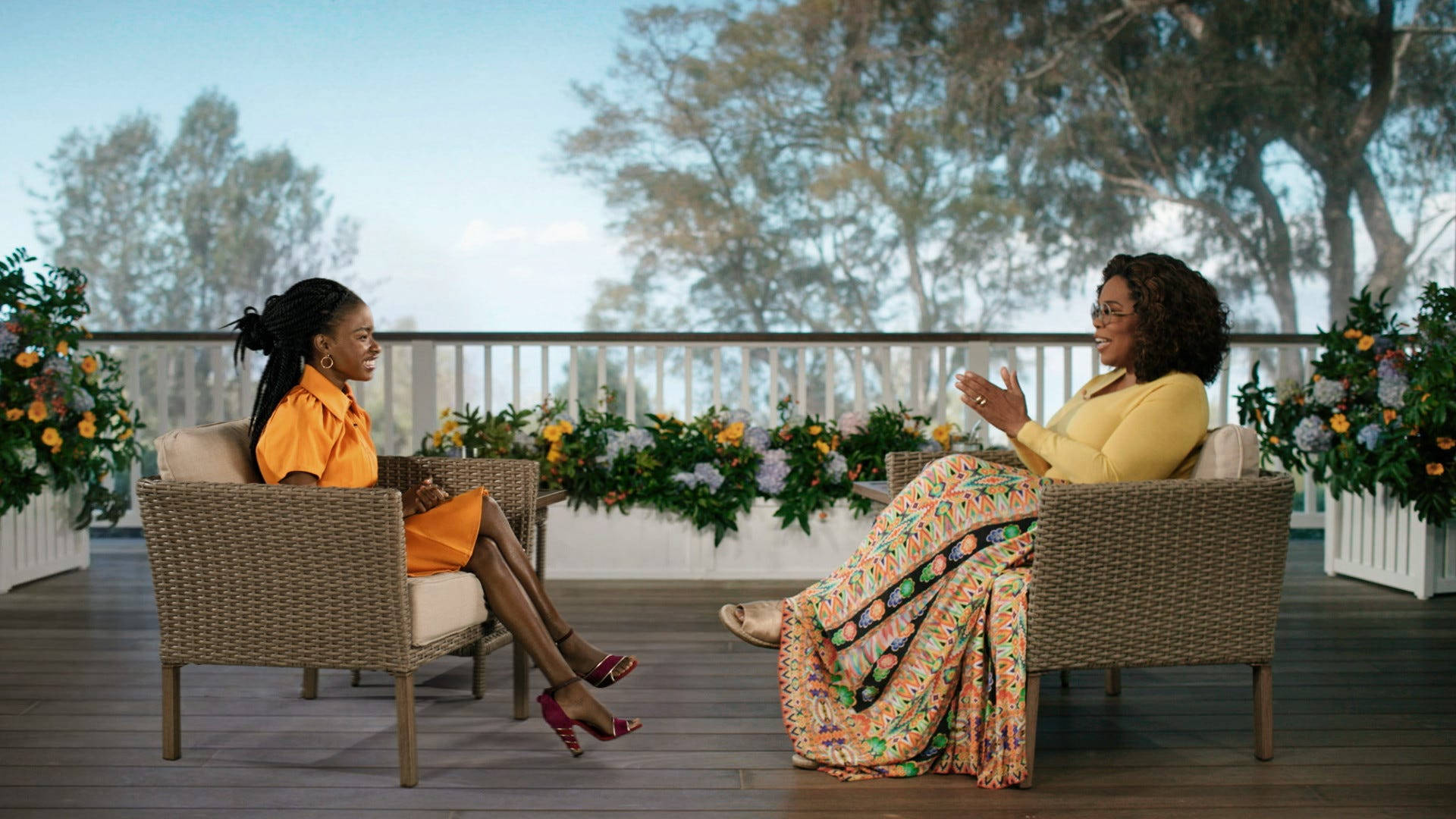 Oprah 桌布 壁紙 — 兩位穿著鮮豔服裝的女性在一個裝飾著茂盛花卉和樹木的木質甲板上，在柔和日光下溫暖交談。