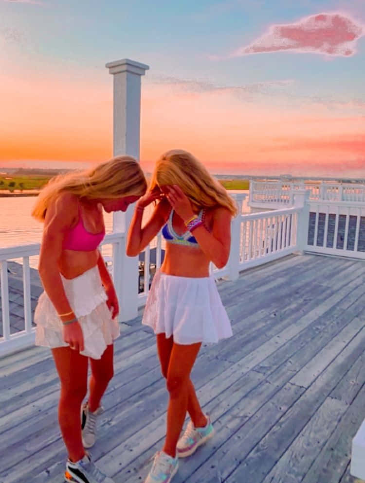 Best Friend Aesthetic 桌布 壁紙 — 兩位穿著鮮豔比基尼和白色裙子的女士在日落時分的木製碼頭上大笑，天空呈現粉紅色和橙色。