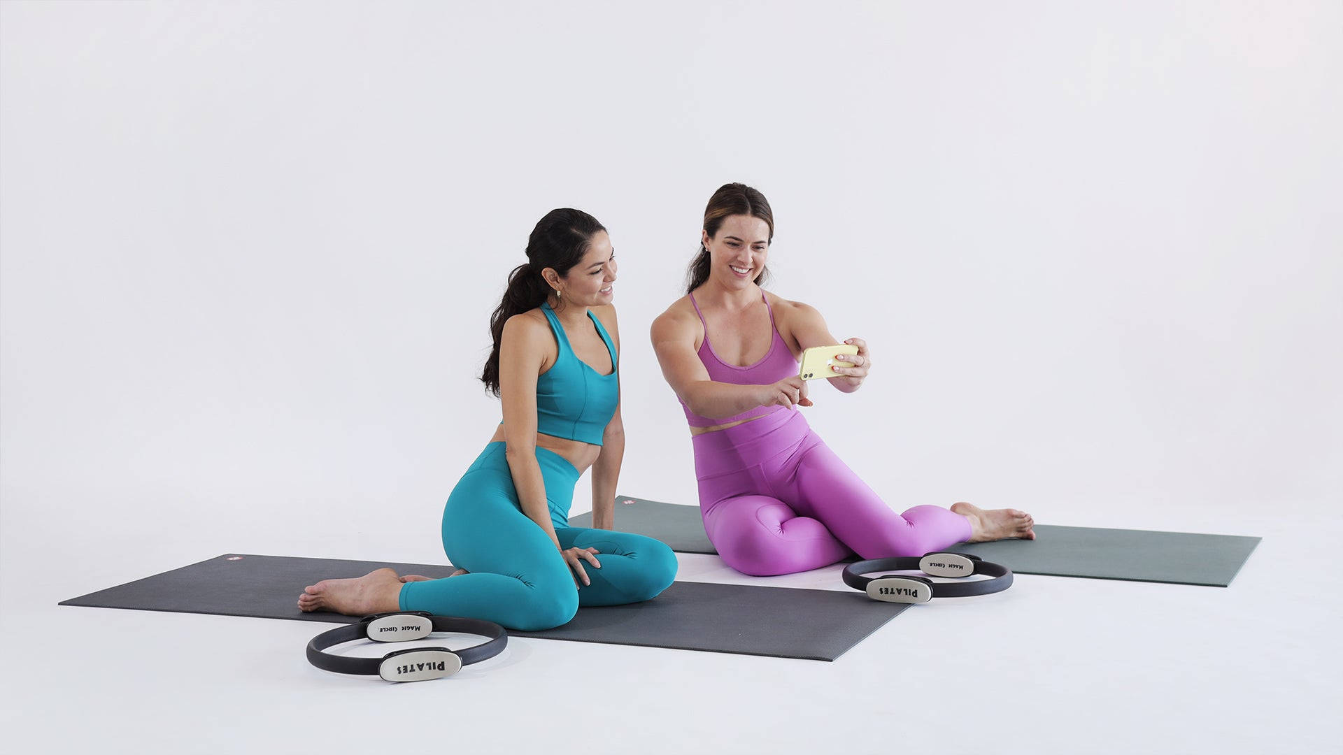 Exercise 桌布 壁紙 — 兩位穿著鮮豔運動服的女士坐在瑜珈墊上，旁邊放著皮拉提斯環，在潔白的背景下自拍。