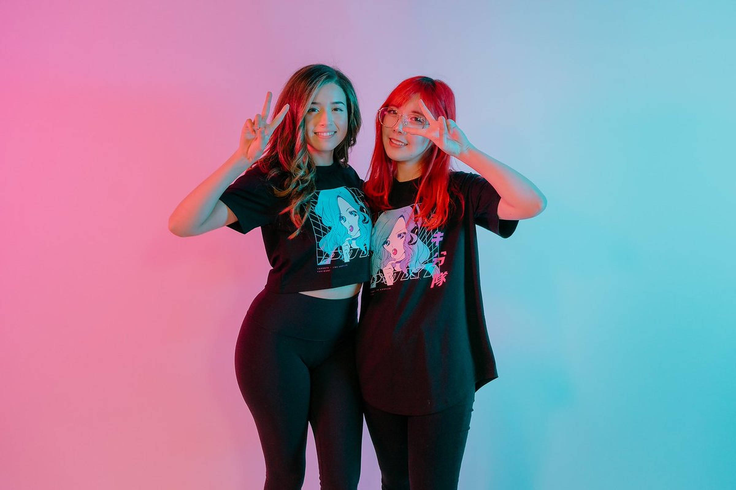 Pokimane 桌布 壁紙 — 兩位笑容滿面的女性，擁有充滿活力的粉紅色和藍色頭髮，在漸變霓虹牆前愉快地擺姿勢，做出和平手勢。