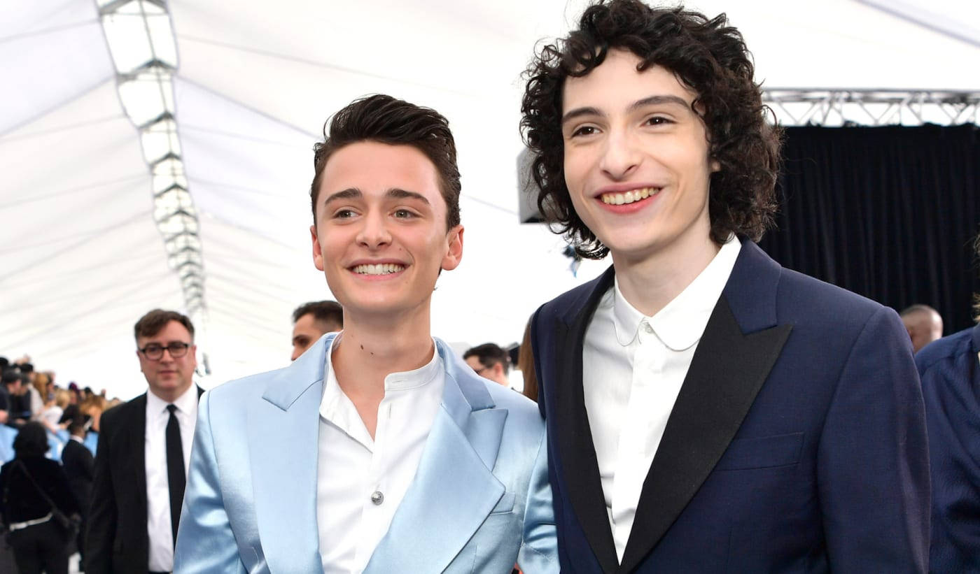finn wolfhard 桌布 壁紙 — 兩位笑容滿面的年輕男士身穿時尚西裝，在白色帳篷下的明亮戶外活動中一起擺姿勢。