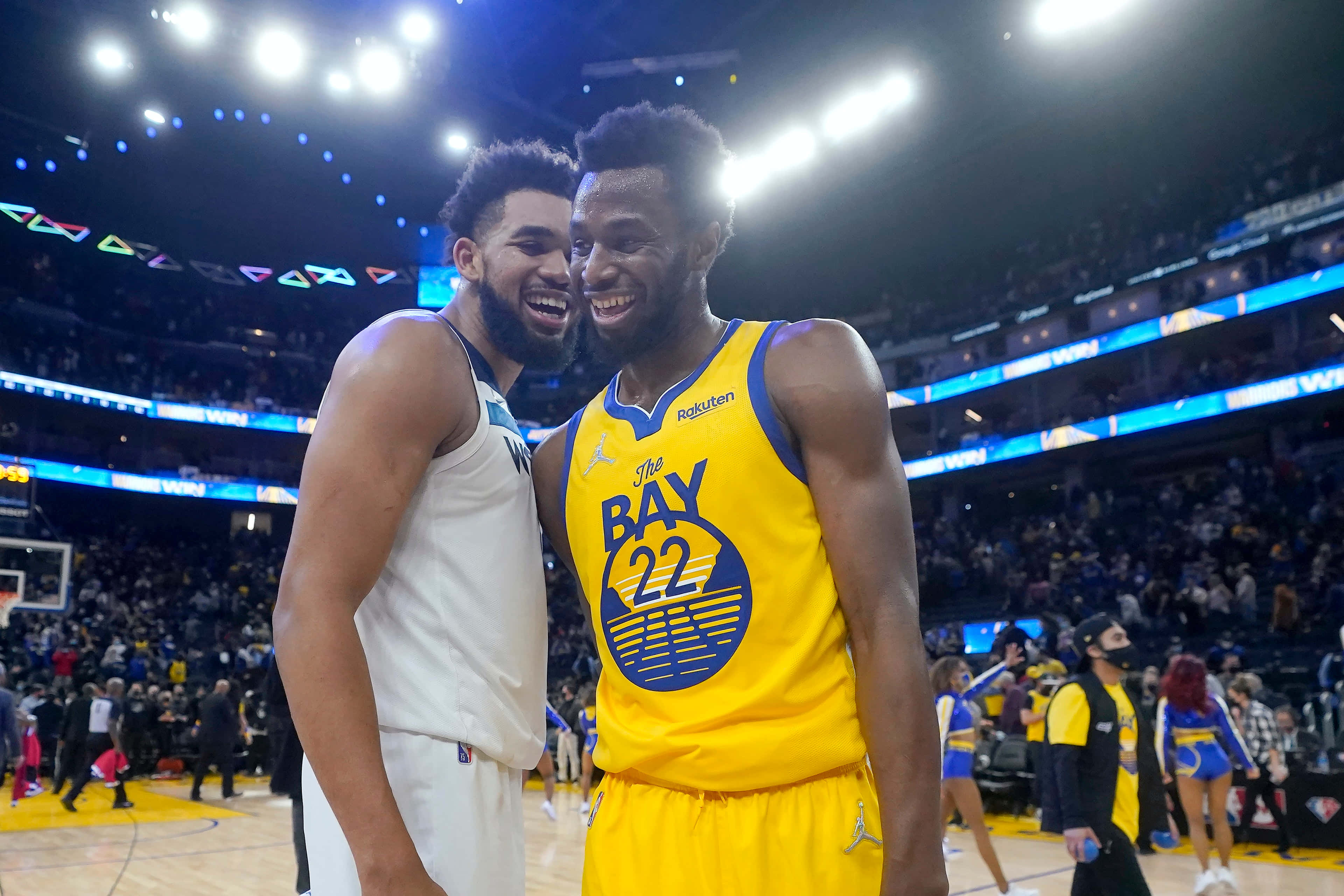 Andrew Wiggins 桌布 壁紙 — 兩位籃球運動員在燈光明亮的球場上與背景中歡呼的球迷一起開懷大笑。
