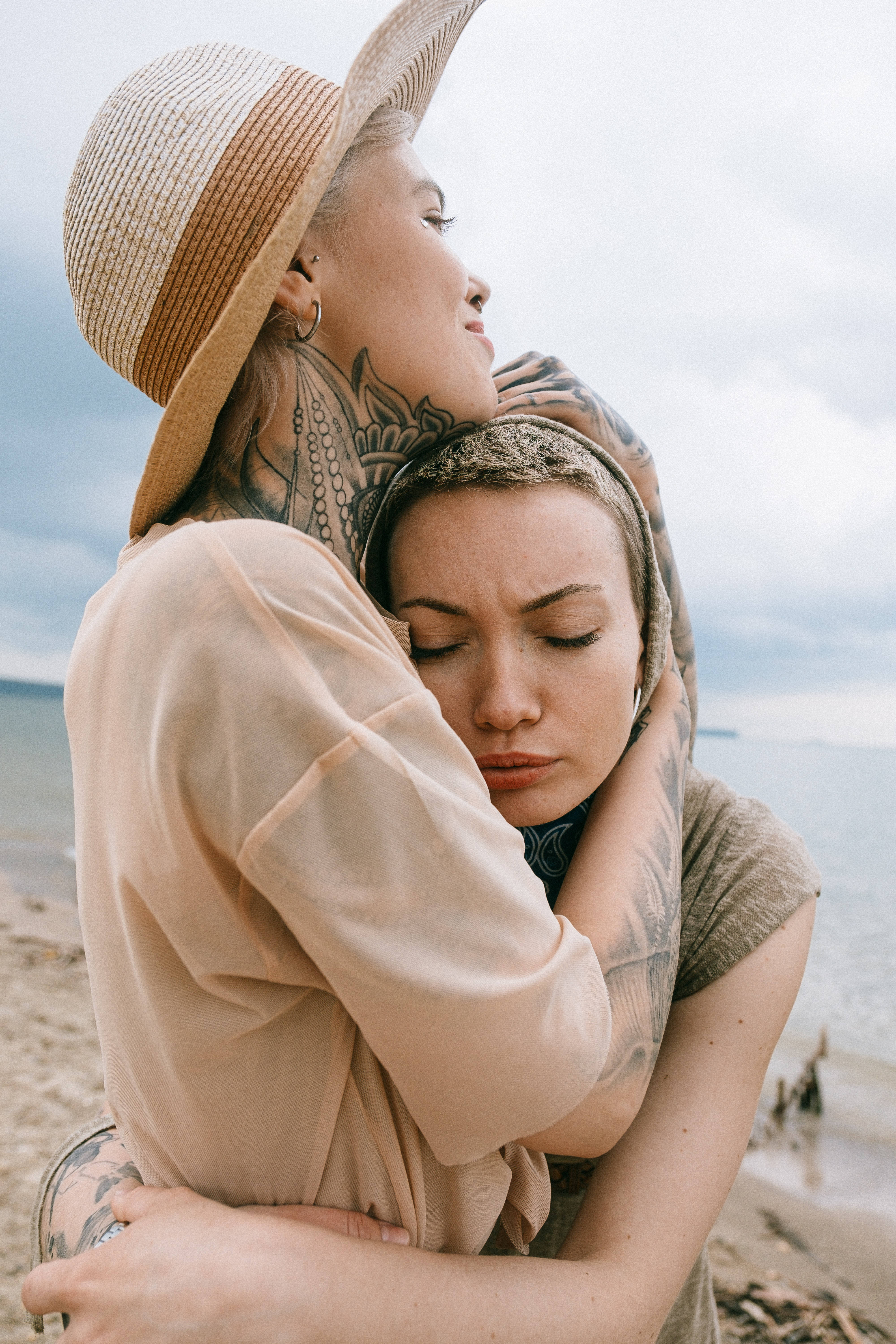 Hugging 桌布 壁紙 — 兩位紋身女士在陰天的海灘上溫柔擁抱，背景是海浪。