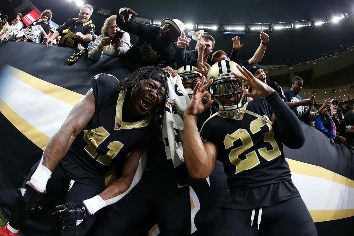 Alvin Kamara 桌布 壁紙 — 兩位紐奧良聖徒隊美式足球員在球場燈光下與高舉巨型球隊旗幟的球迷一同勝利慶祝。