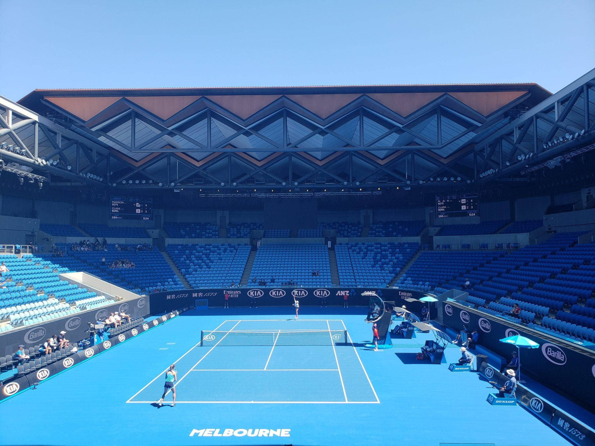 Australian Open 桌布 壁紙 — 兩位網球運動員在晴朗天空下，於一個擁有幾何屋頂建築的現代化體育館內的鮮豔藍色球場上比賽。
