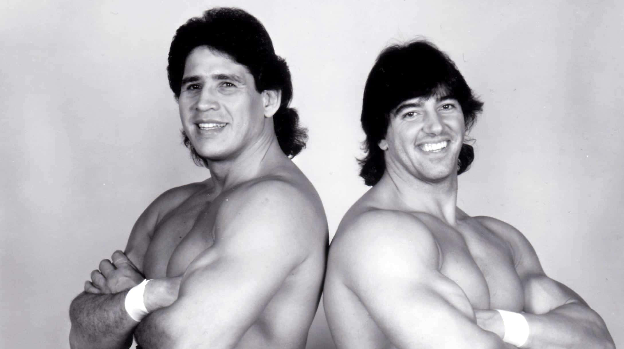 WWE 桌布 壁紙 — 兩位肌肉發達、頭髮長及肩膀的男士背對背站立，在黑白工作室肖像中自信地微笑。